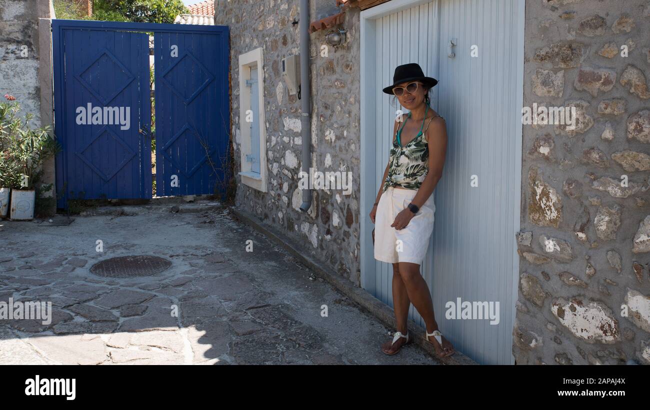 Asian Woman in sunglasses and hat standing in front of traditional Greek house with blue shutters and doors in village of Eressos on islands of Lesvos Stock Photo
