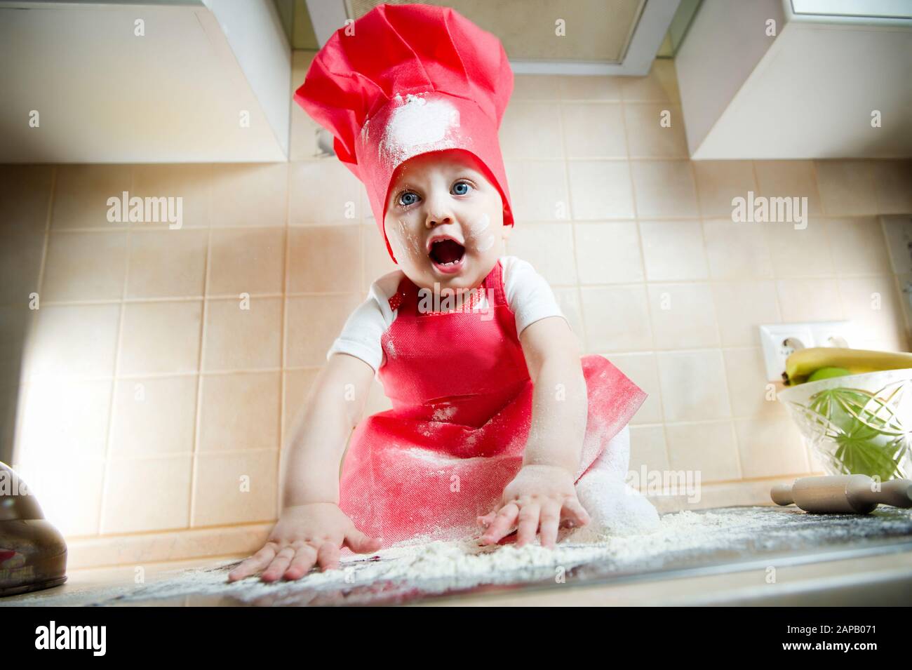 little girl in a cook suit is ethnically screaming, sitting on the stove and playing with flour. Little cook in a suit Stock Photo
