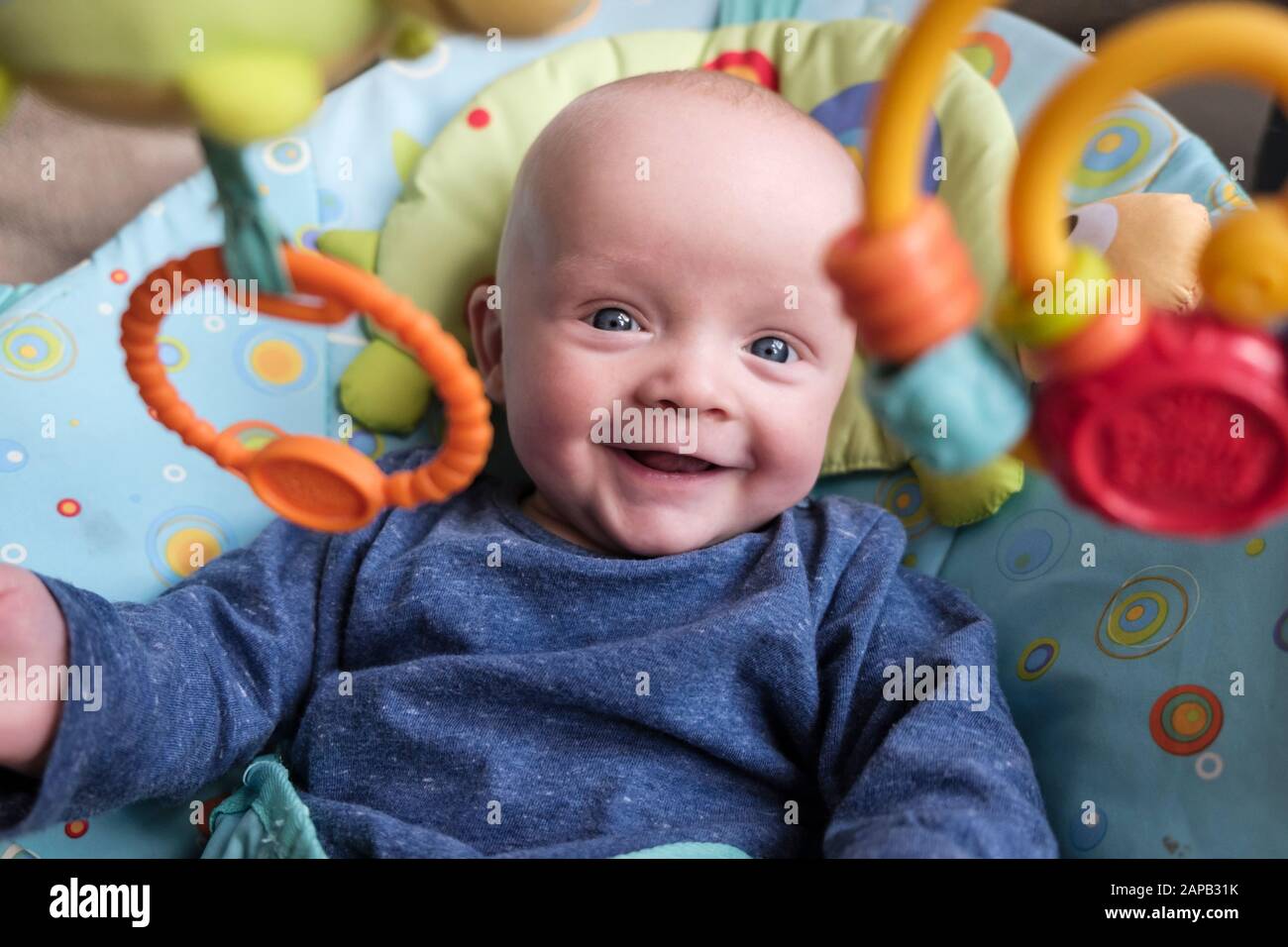 A happy baby boy age five months in an activity chair smiling and looking forwards at hanging toys. England, UK, Britain Stock Photo