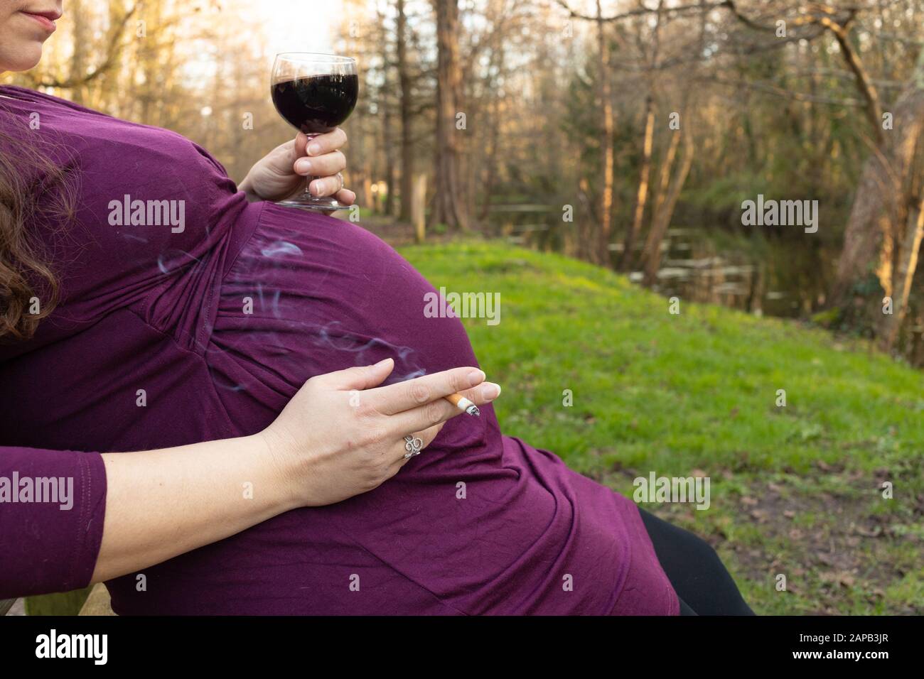 Pregnant woman sitting on a bench, drinking red wine and smoking cigarette in a park. Stock Photo