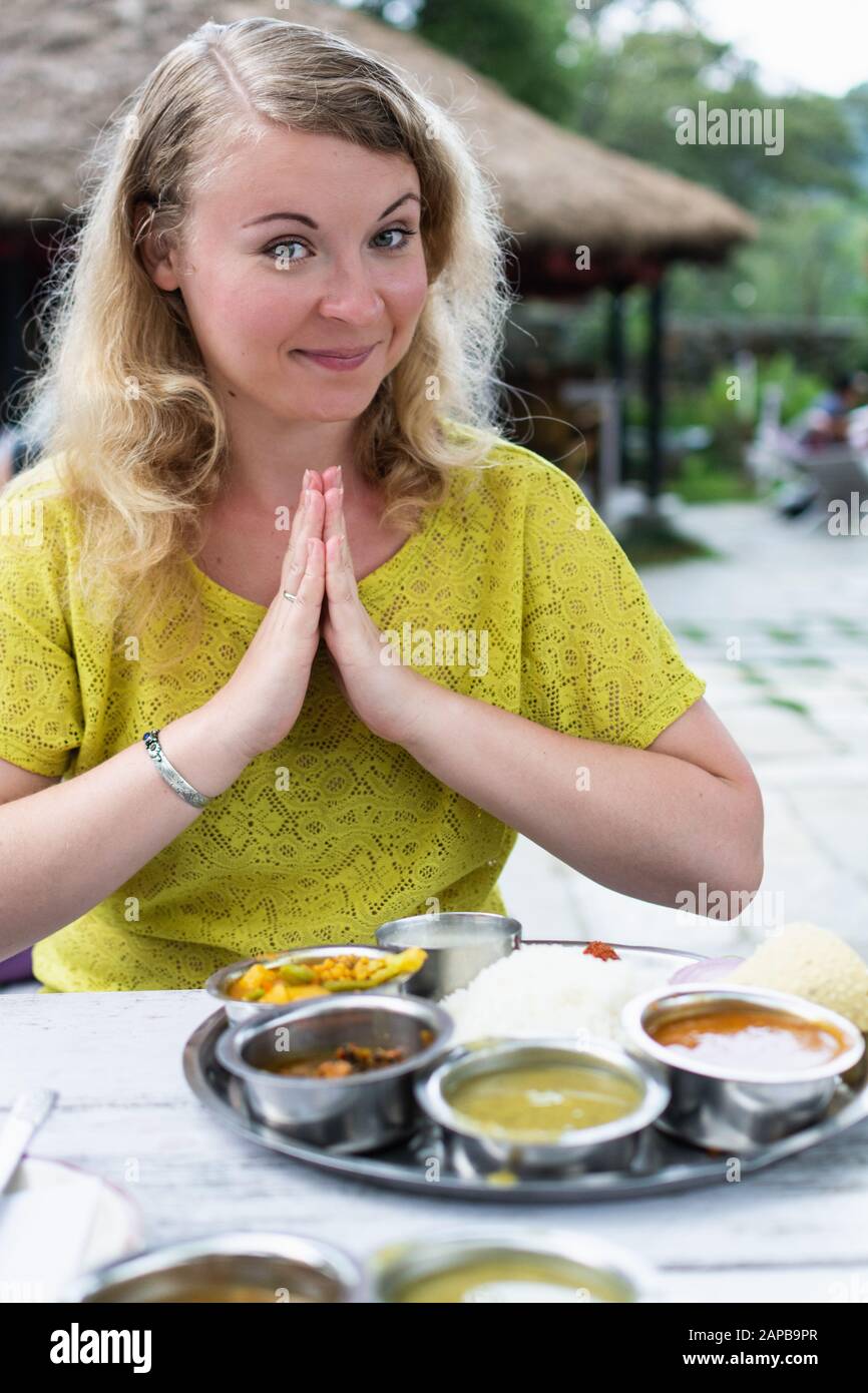 Blonde woman at the table in Indian restaurant with dal bhat served with steamed rice and lentil soup, pressing hands together with a smile to greet N Stock Photo