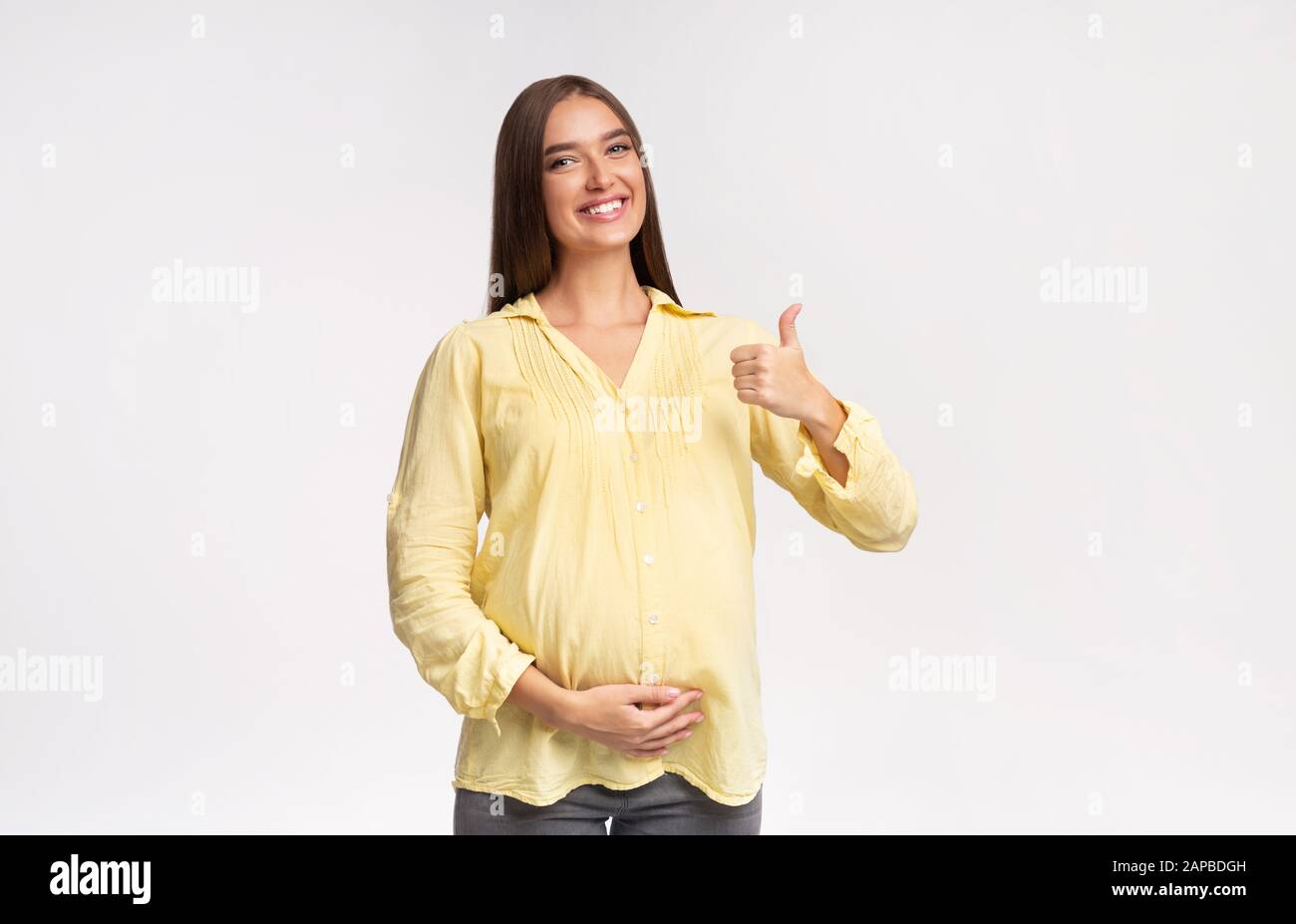Joyful Pregnant Girl Gesturing Thumbs-Up Standing On White Studio Background Stock Photo