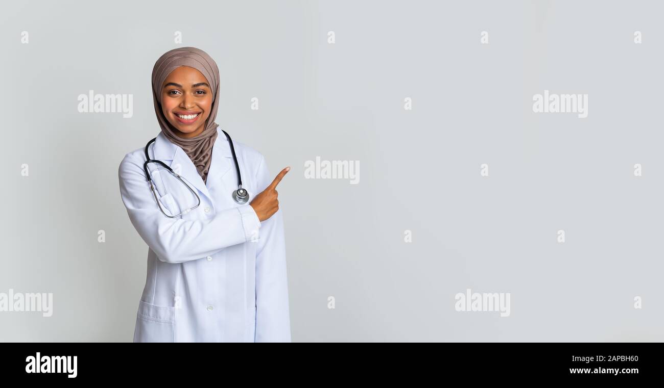 Positive black female therapist in hijab and white coat pointing aside Stock Photo