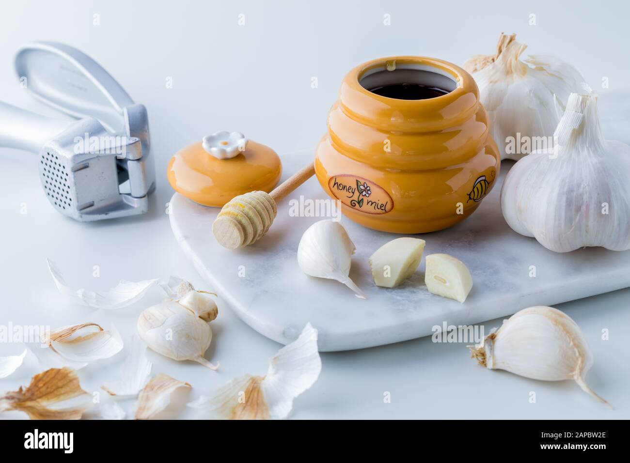 A close up of a honey pot and dipper surrounded by garlic cloves and a garlic press. Honey