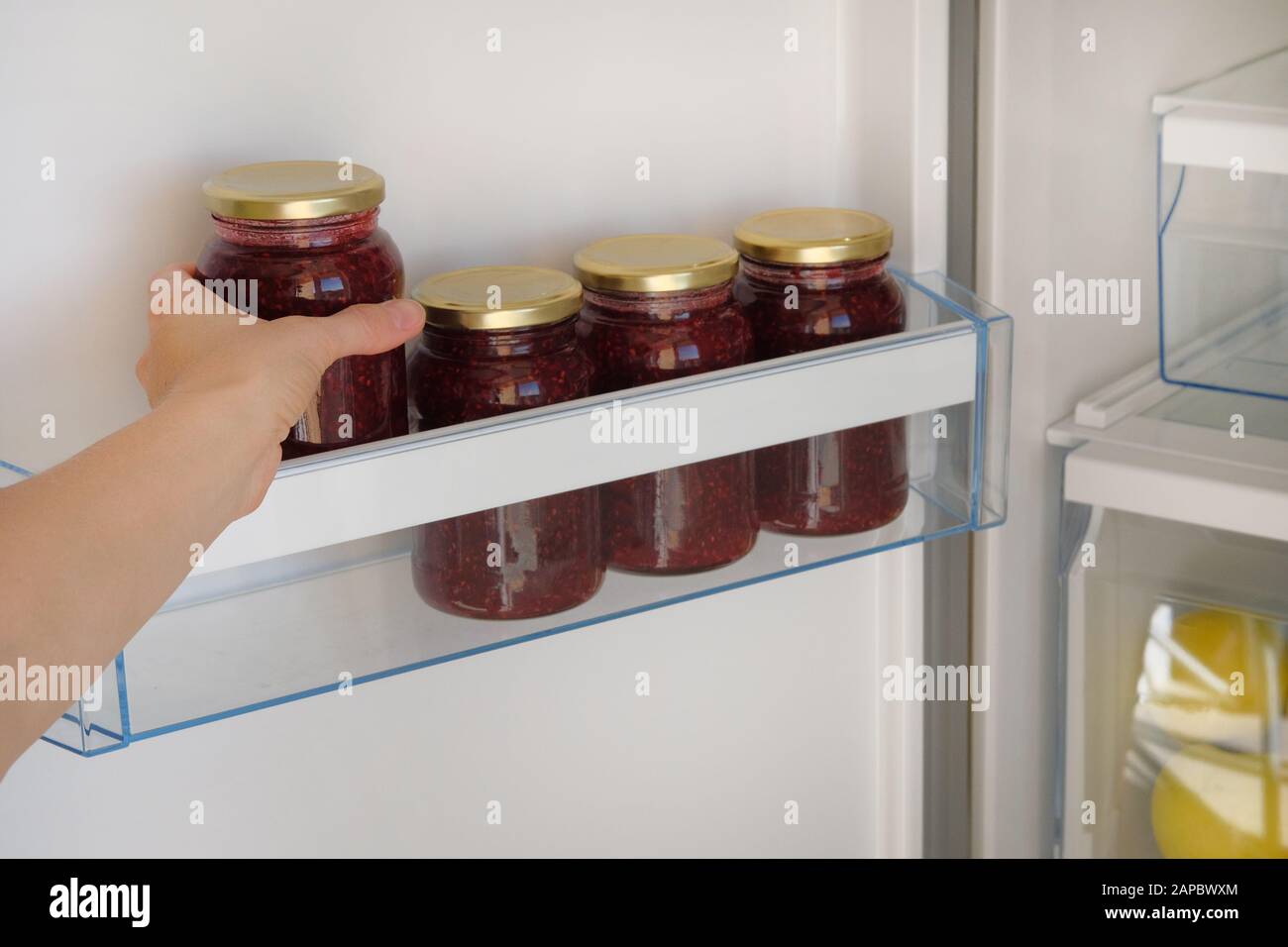 Glass Jars with Raspberry Jam on shelf in fridge. Female hand with a jar of red homemade jam. Fermented healthy vegetarian foods. Stock Photo