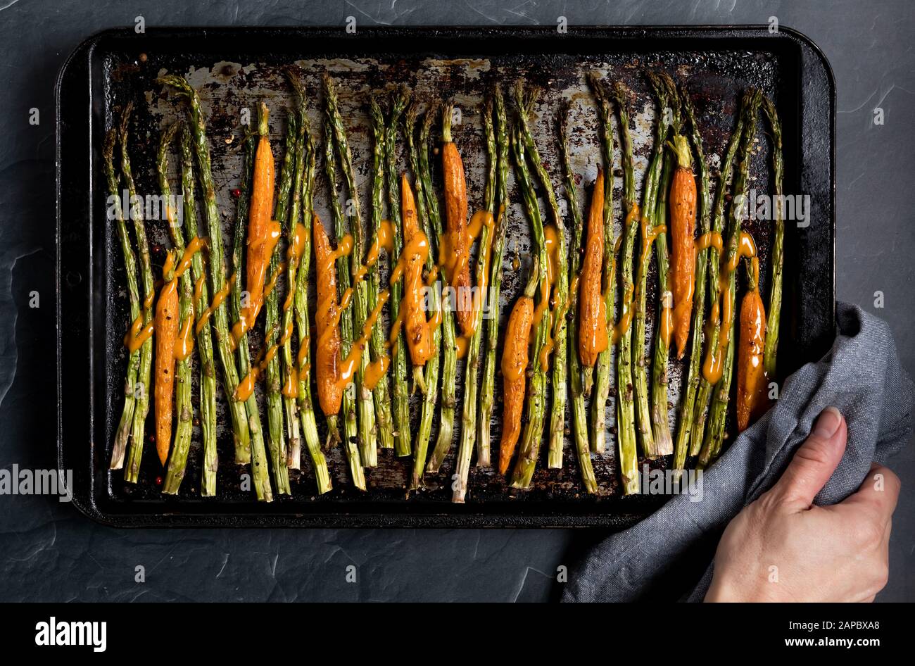 A top down view of a baking sheet of oven roasted asparagus and baby carrots and drizzled with sauce. Stock Photo