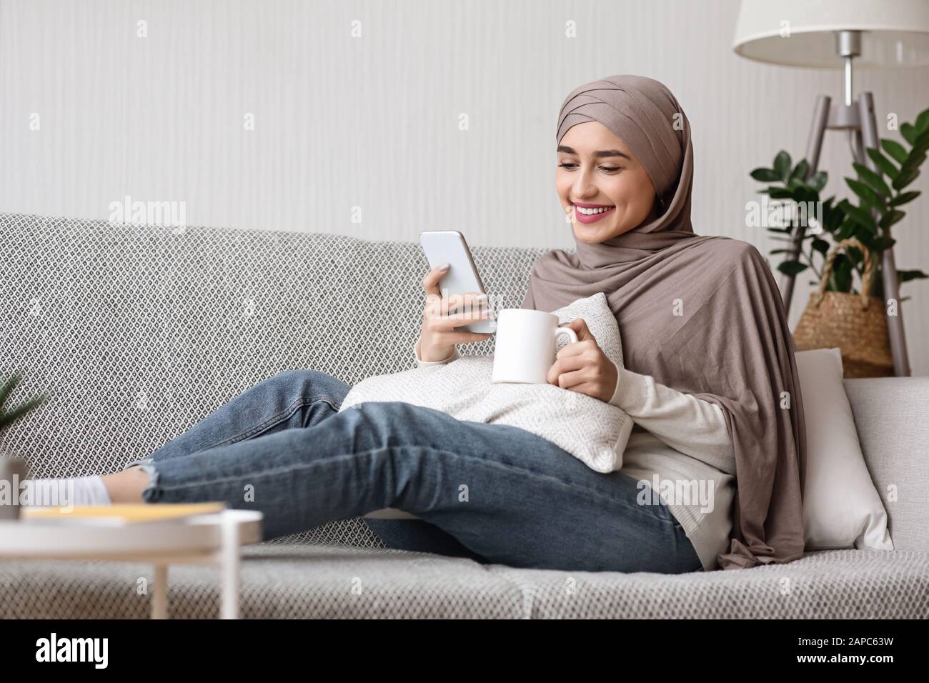 Muslim girl in hijab relaxing on sofa with smartphone and coffee Stock ...