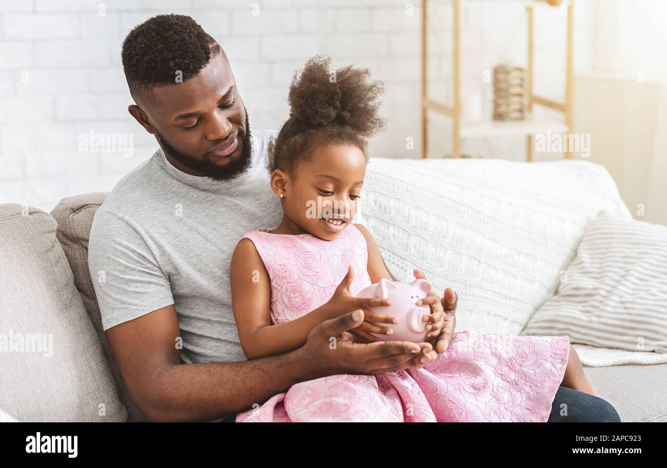 Young afro father showing little daughter piggy bank Stock Photo - Alamy
