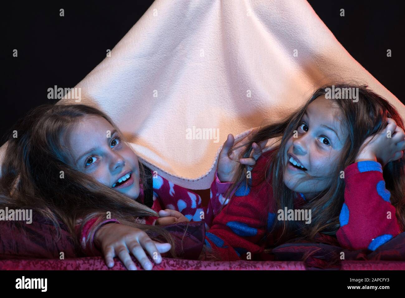 Pyjamas party for children. Girl friends under blanket pulling on each  others hair. Girls with concentrated faces. Kids wearing red jammies in bed  on black background. Children and PJs party concept Stock