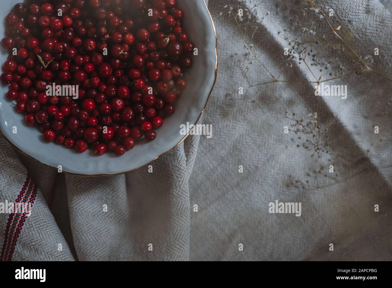 top down view on lingonberries in a bowl on linen tablecloth and some fragile flowers, inside, rustic style with grain Stock Photo