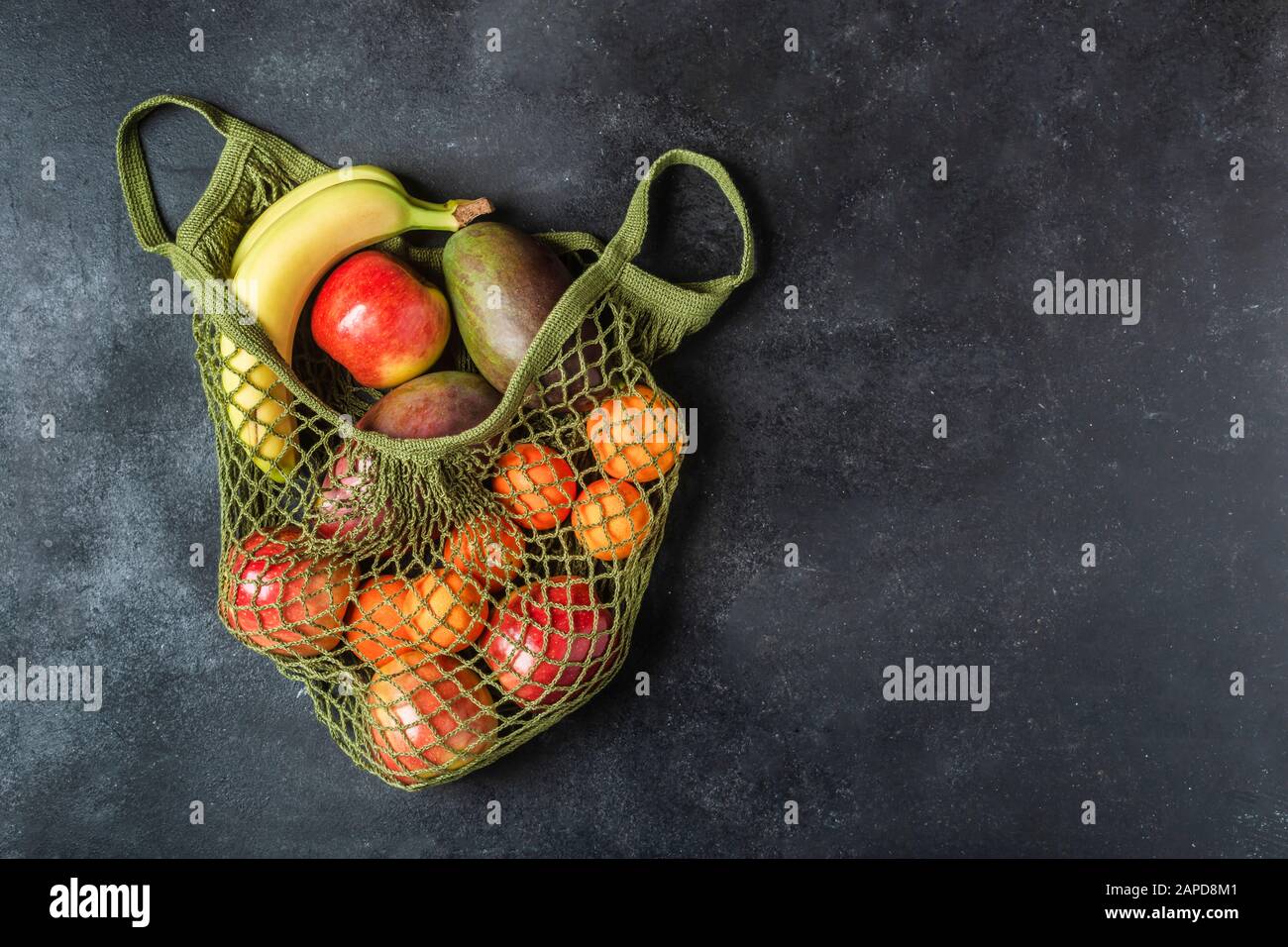 Fresh fruit in a green string bag on a black background. Bananas, apples, oranges, and mangoes. Stock Photo