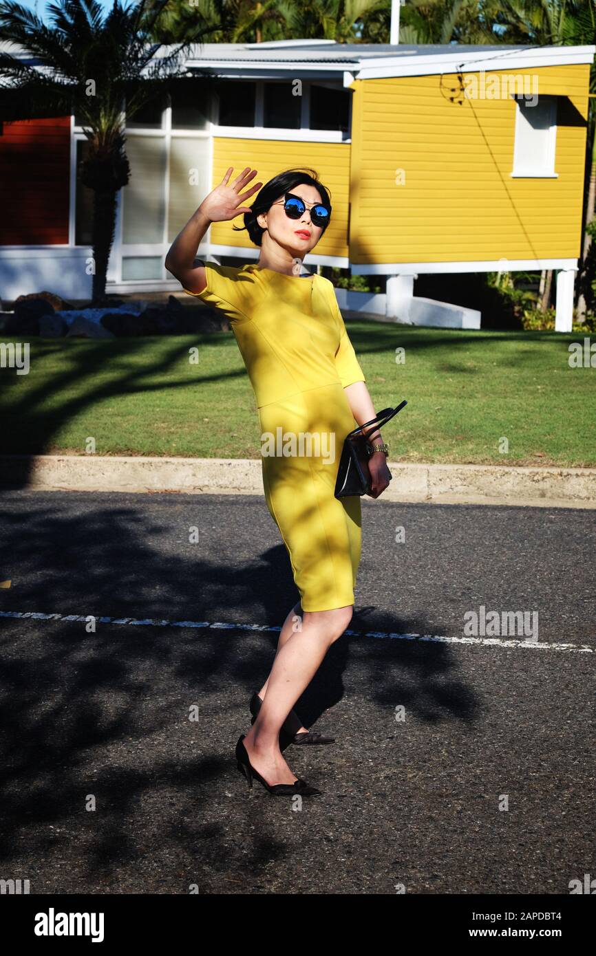A glamourous woman in a sixties-inspired fashion photo shoot, Carina Brisbane with a mid-century modern house, yellow dress, blue sunglasses Stock Photo