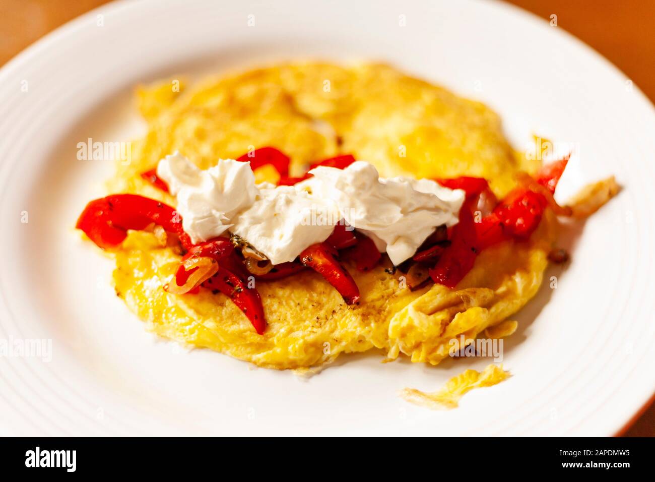 An omelette with sauteed bell peppers, shallots, and sour cream is plated on a white dish with a wooden table as background. Stock Photo