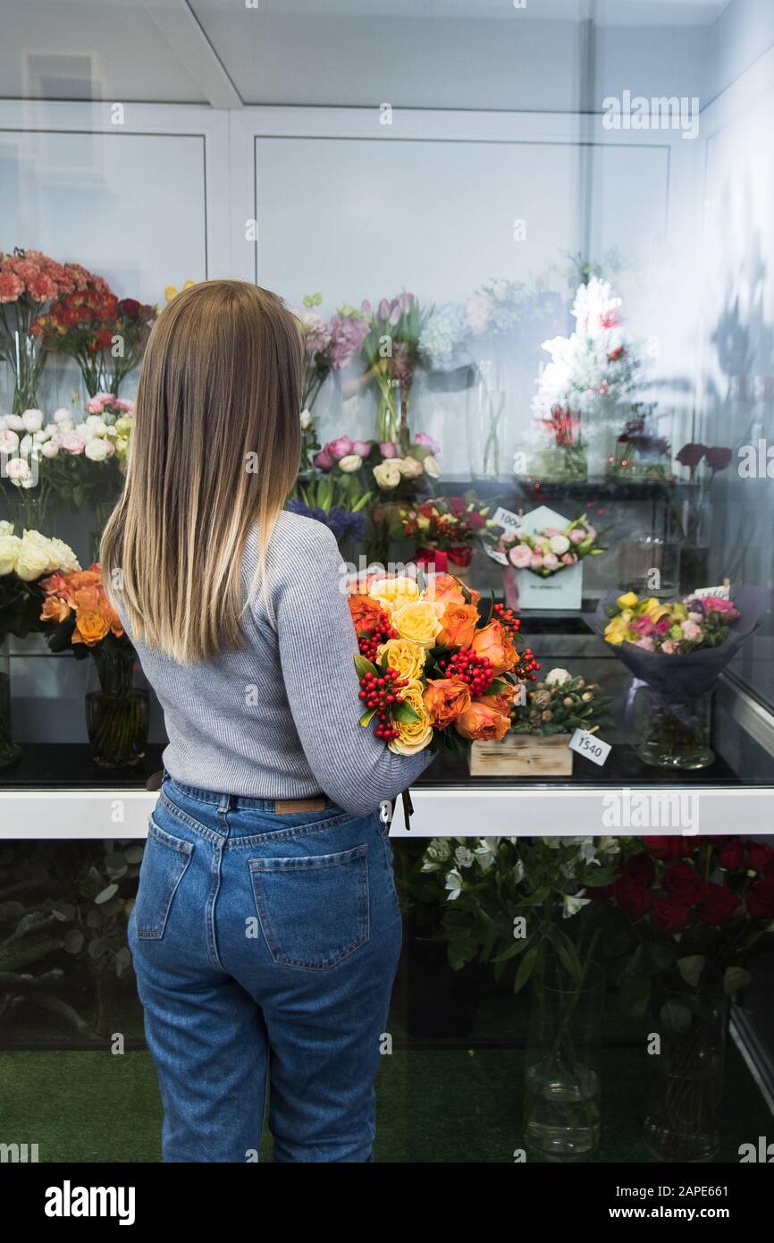 The girl holds a beautiful bouquet of orange and red flowers, roses, next to the flower counter. Lovely woman chooses a bouquet in a florist shop. Stock Photo