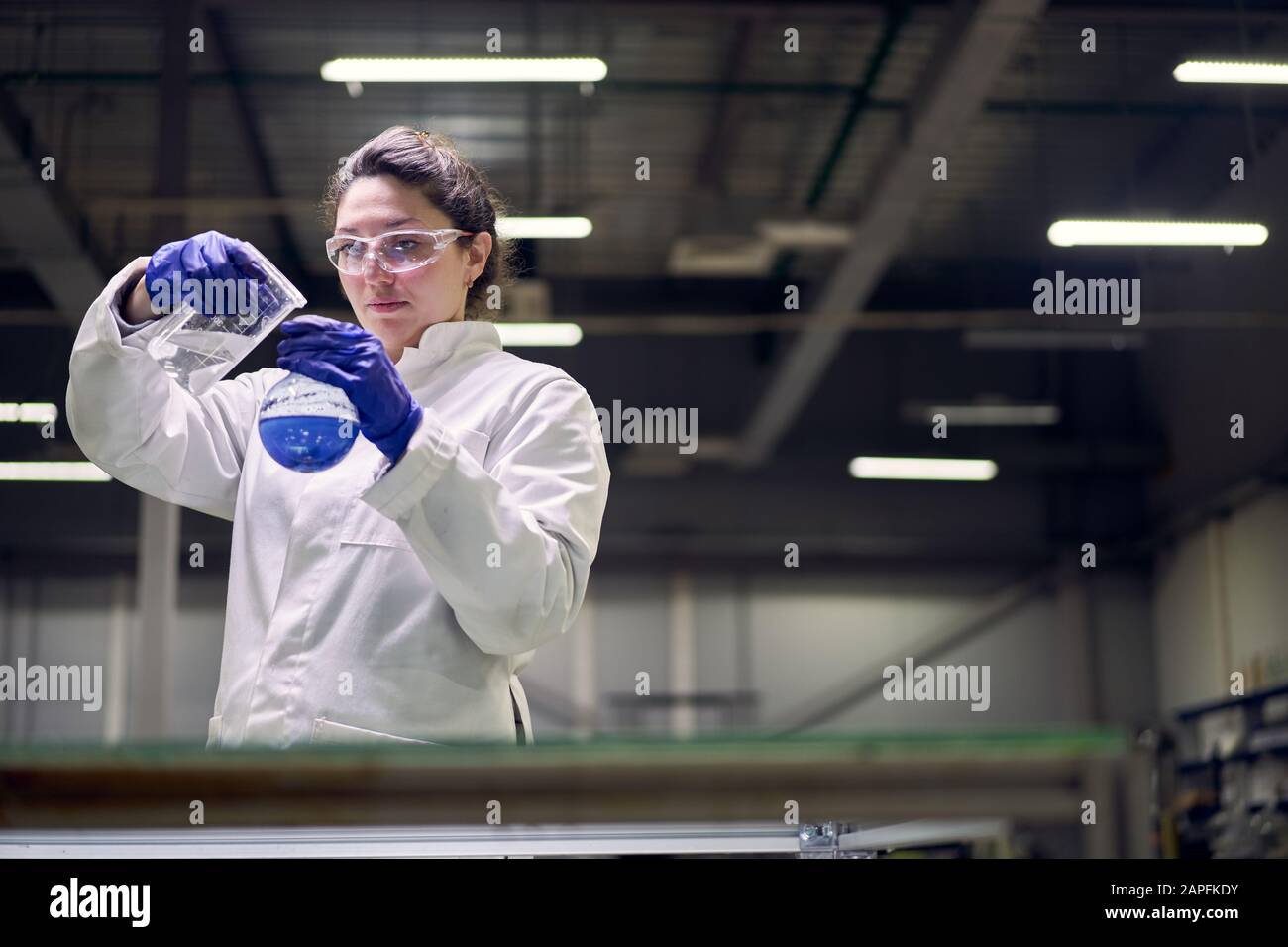 Young lab woman in glasses and white coat with flask with blue liquid ...