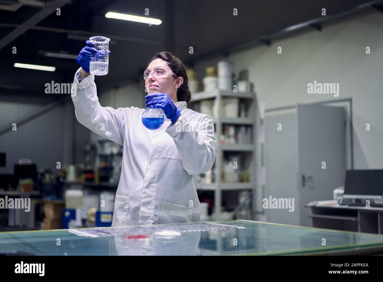 Brunette lab girl in glasses and white coat with flask with blue liquid ...