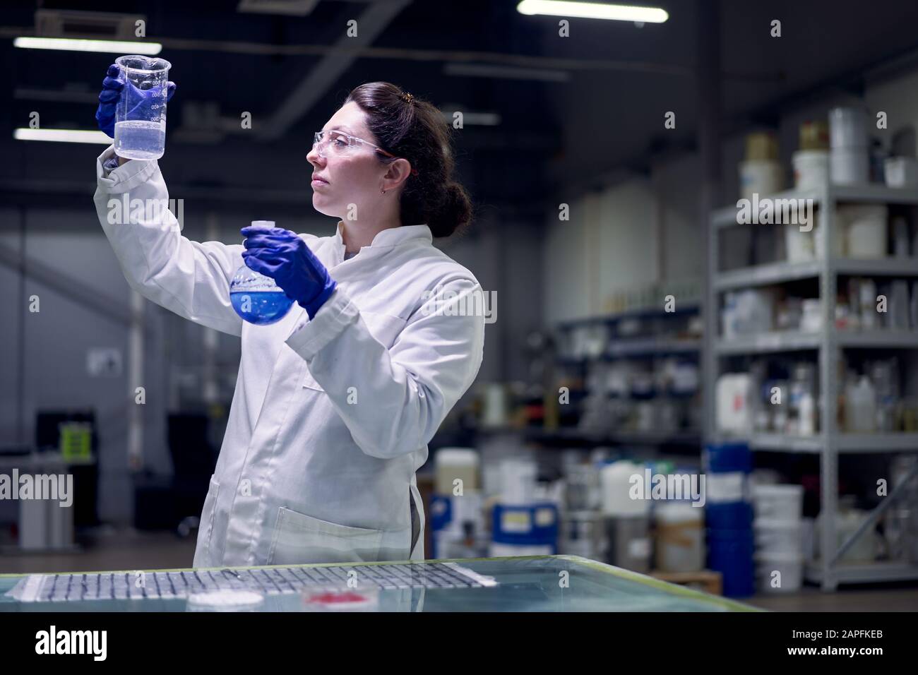 Young lab woman in glasses and white coat with flask with blue liquid ...