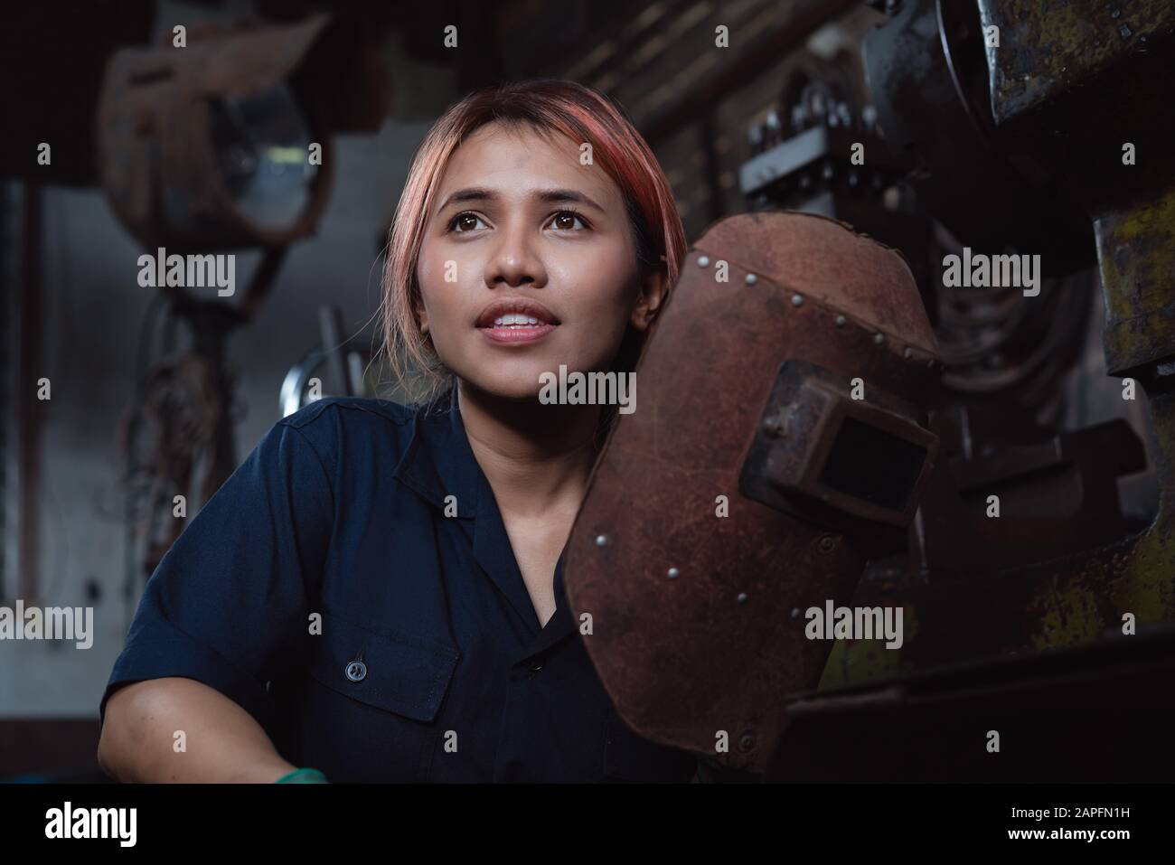 Diverse female industrial engineer holding welding helmet after work shift - Young Asian factory metal worker taking a break - Hispanic apprentice woman, learning new skills on internship training Stock Photo