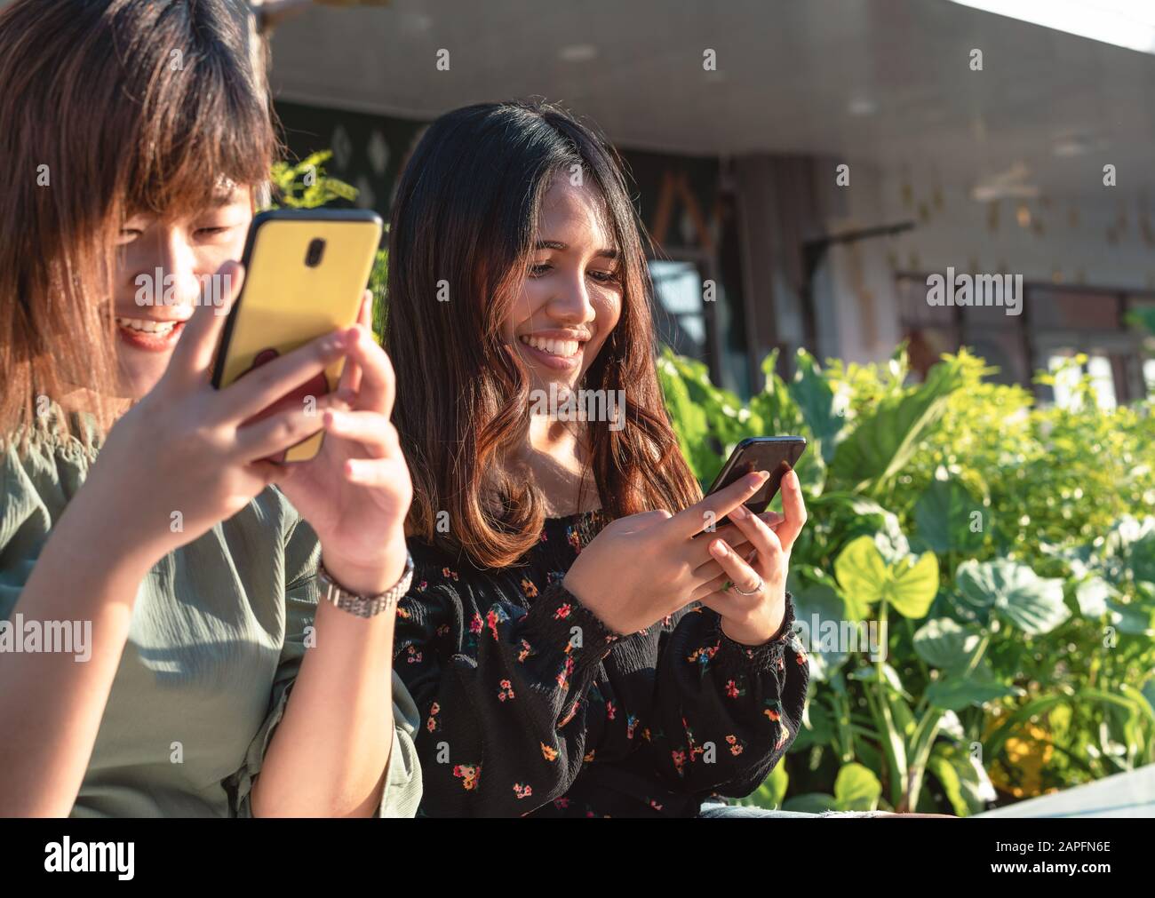Young millennial Asian friends hanging out outdoors instant messaging on smartphones - Happy smiling hipster girls chatting on social media using mobile phone - Leisure, lifestyle and pals concept Stock Photo