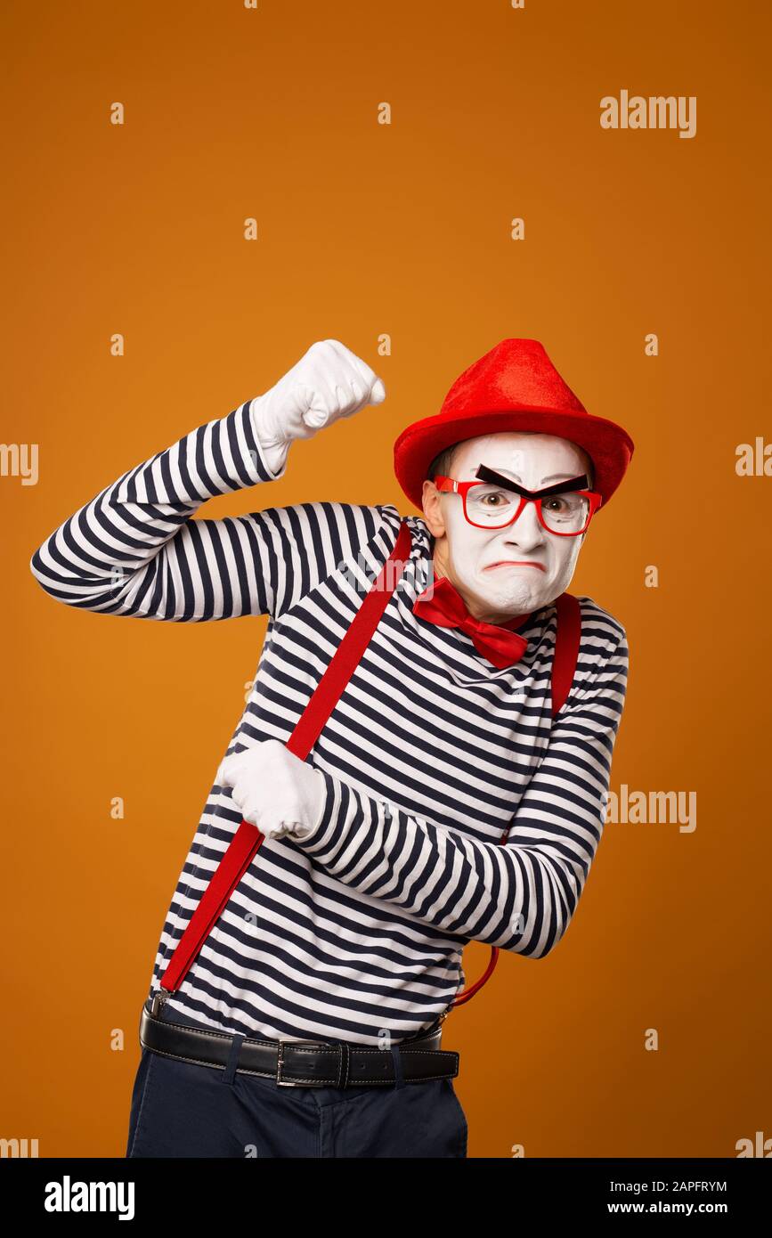 Sad mime with white face in red hat and striped t-shirt on blank orange background in studio Stock Photo