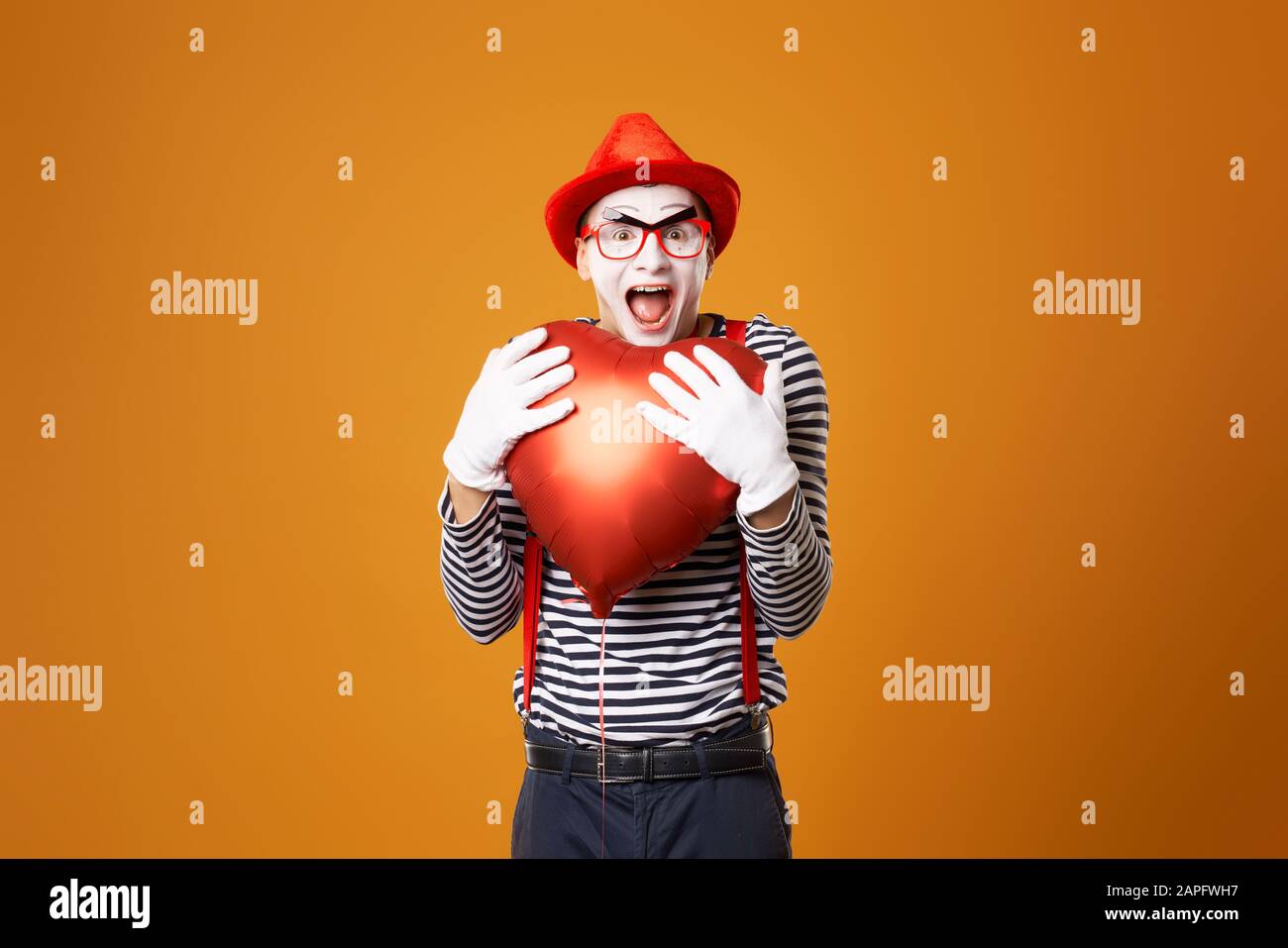 Sad clown mime in red hat and vest holds heart ball in his hands on empty orange background Stock Photo