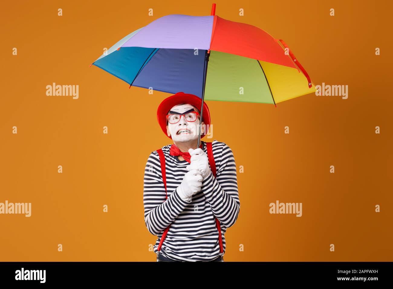 Dissatisfied mime man with multi-colored umbrella isolated on blank orange background in studio Stock Photo