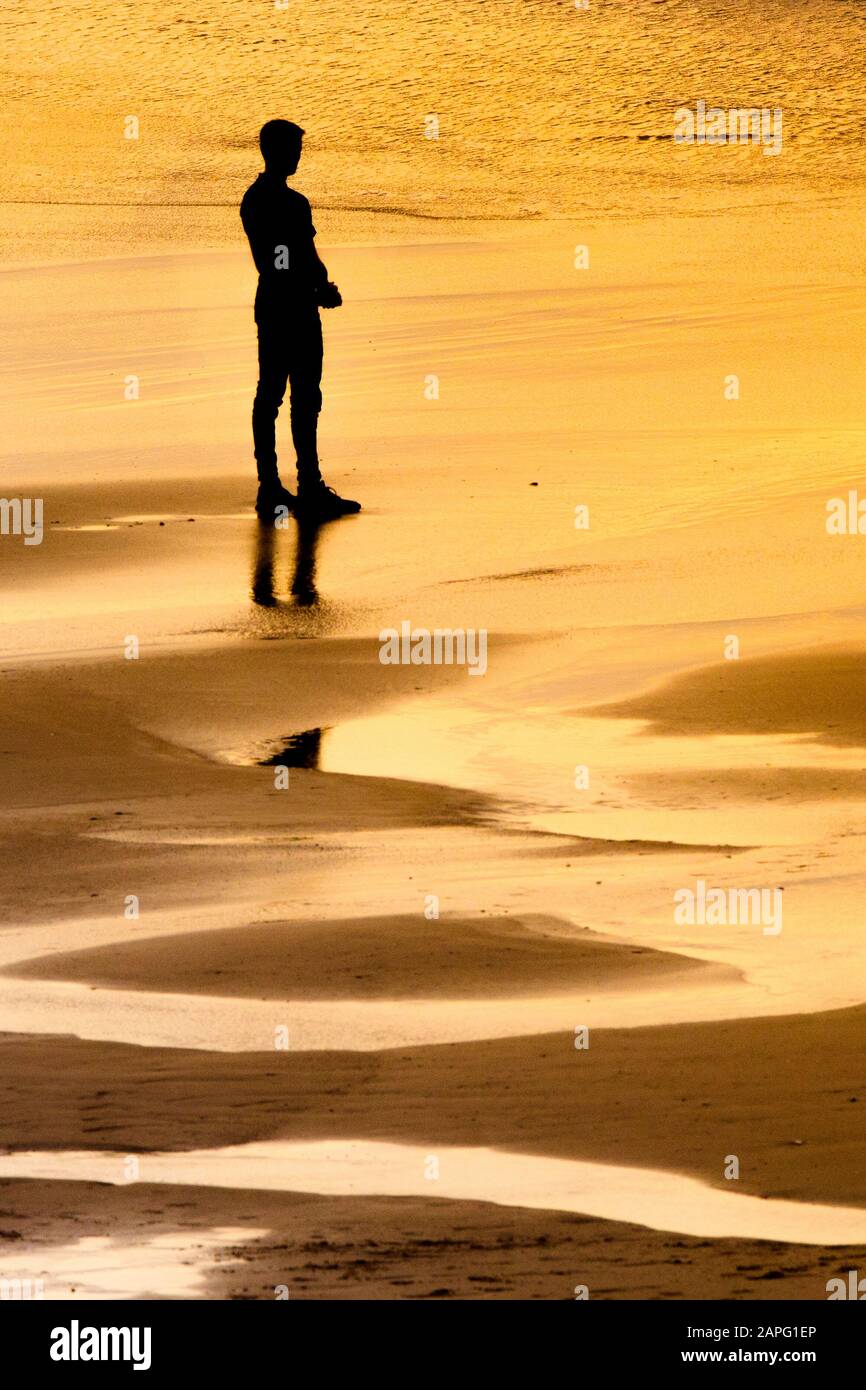 A lone male figure silhouetted by the setting sun as he stands looking out to sea at Fistral beach in Newquay in Cornwall. Stock Photo