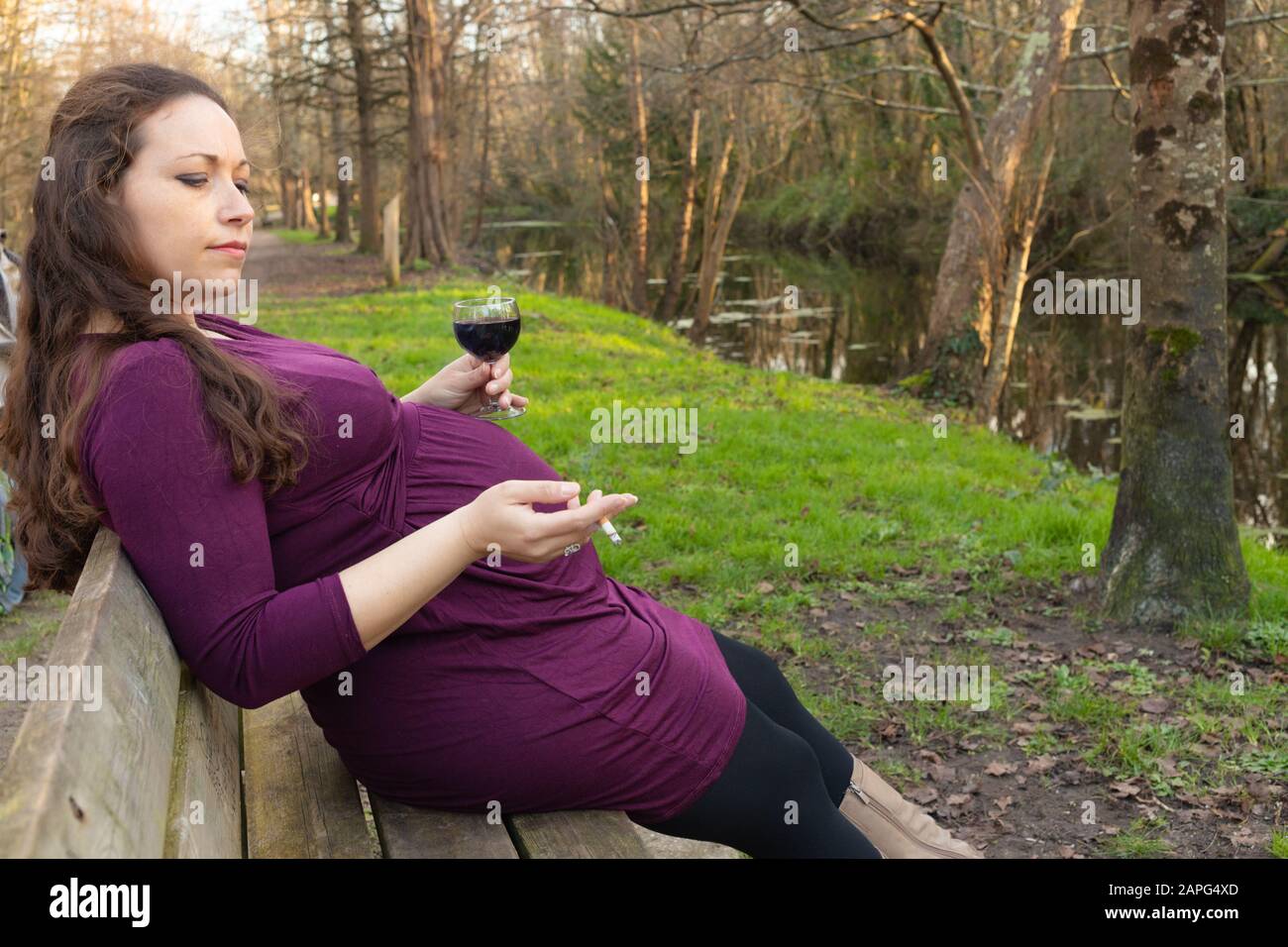 Pregnant woman with alcohol and cigarette in her hands, sitting on the bench in park . Side portrait with copy space Stock Photo