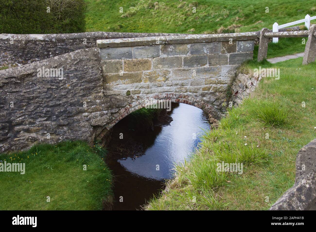 A very small, arched bridge, made from bricks and stone, over a narrow blue stream flowing between high grassy banks. Quaint and cute example of the E Stock Photo