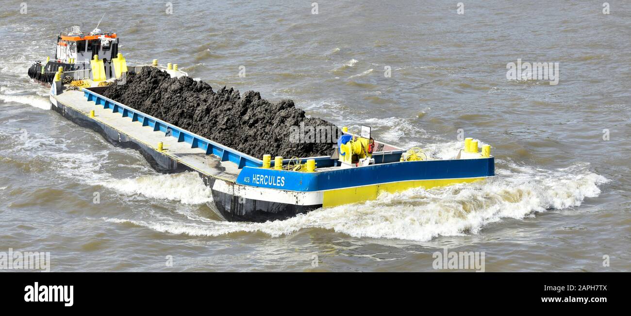 Close up long Hercules barge & load of soil from boring of Thames Tideway Tunnel sewer project tug boat pushing bow wave down river London England UK Stock Photo