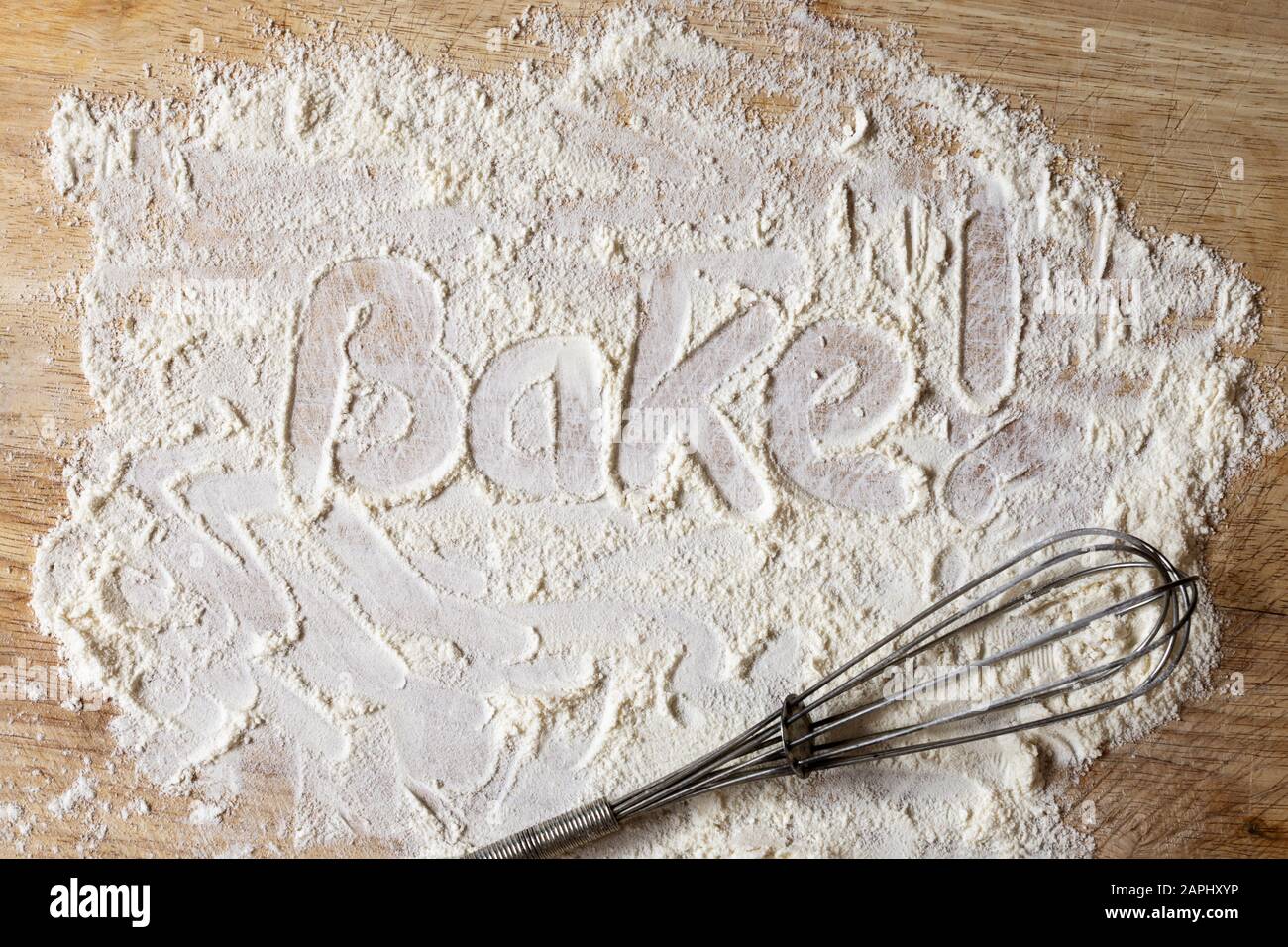 The word 'Bake!' written as an instruction in scattered flour via finger writing on wooden board with retro manual whisk below. Stock Photo