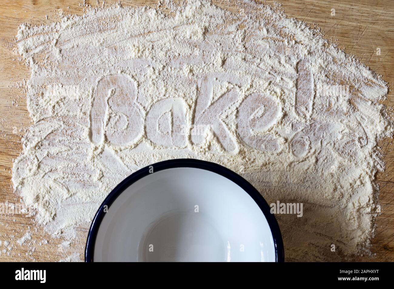The word 'Bake!' handwritten in scattered flour on a wooden chopping board with empty enamel bowl below. Overhead shot. Stock Photo