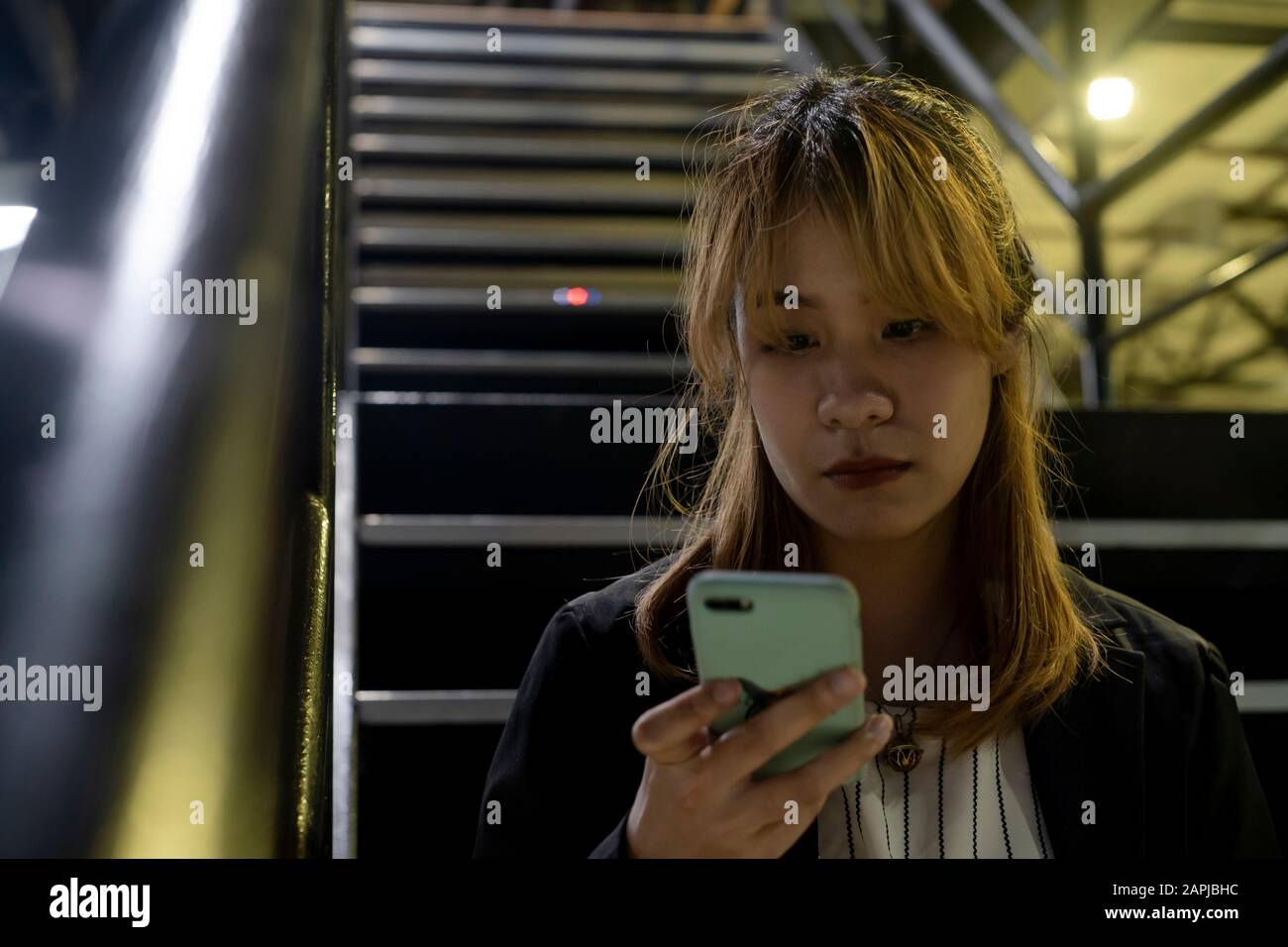 Blond hair woman with fringe in black and white clothes sitting on the stair looking at her smartphone to read the massage from her couple. Stock Photo