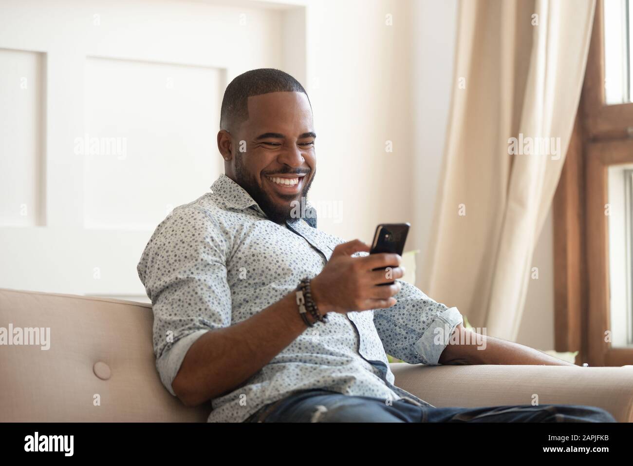 Cheerful african guy sitting on couch spending time using smartphone Stock Photo