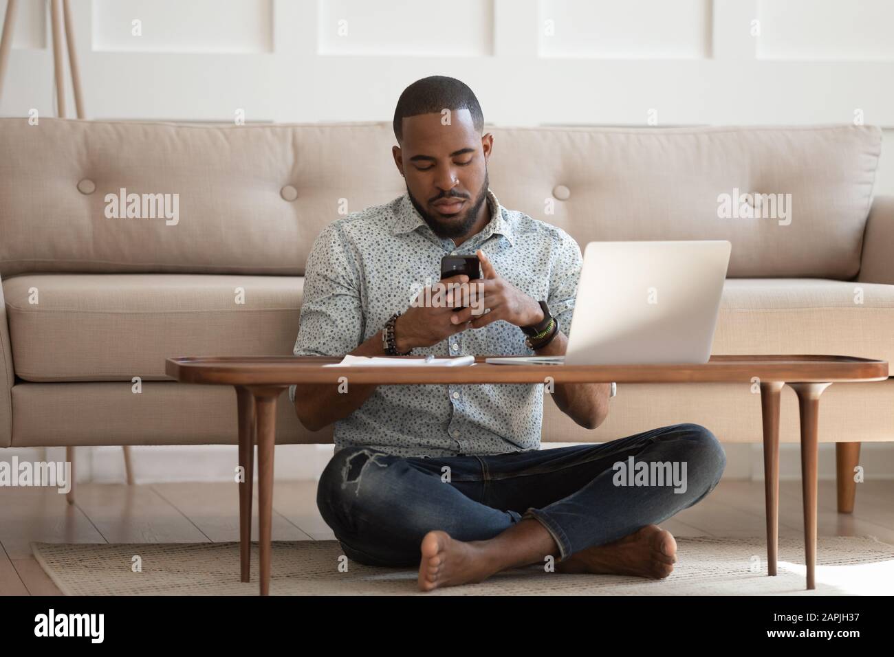 African guy sits cross-legged on carpet using smartphone Stock Photo