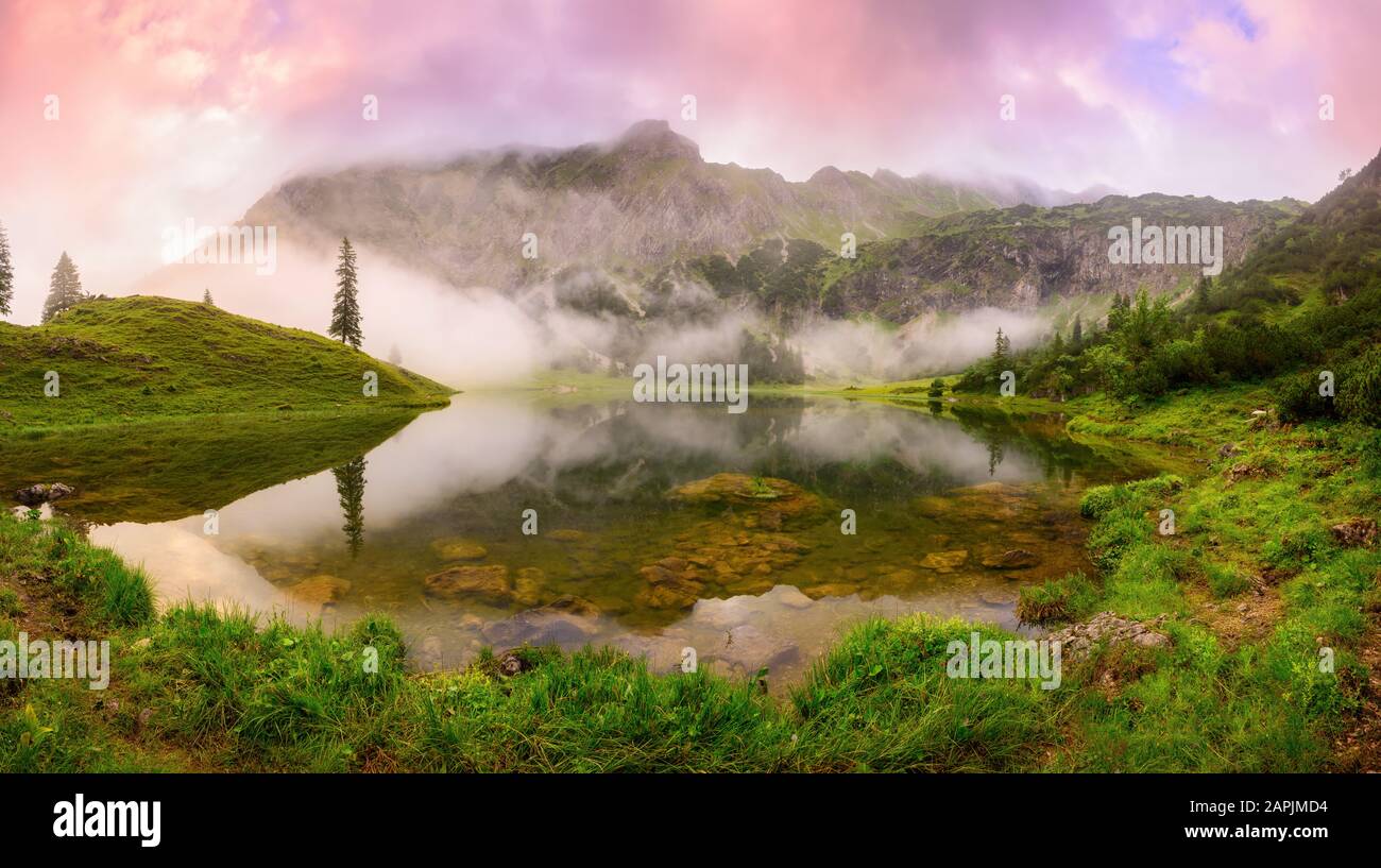 Lake surrounded by mountains at dawn, with colorful clouds and wafts of mist reflected in the water Stock Photo