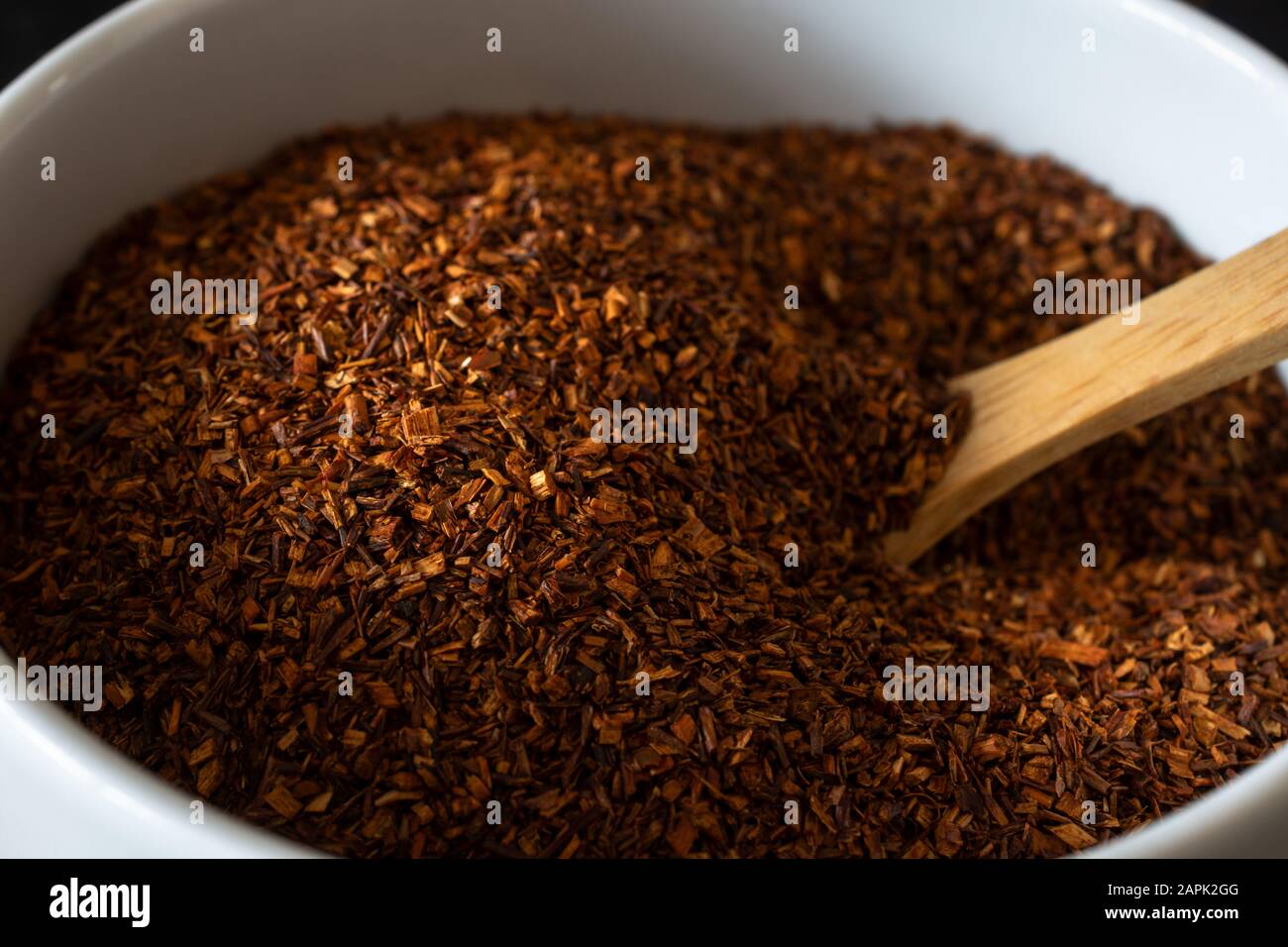 Close up shot of dried Rooibos (Redbush) tea leaves in a white ceramic bowl with wooden spoon. Stock Photo