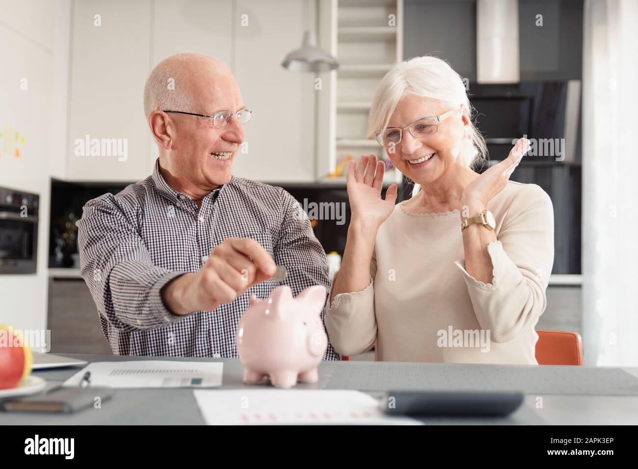 Funny photo of mature couple putting coin into piggy bank at home. Savings, pension plan concept Stock Photo