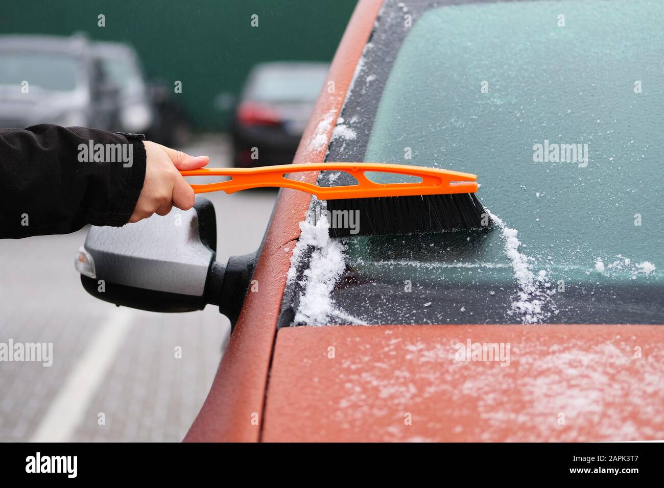 Brush in mans hand. Man clears snow from icy windshield of car. Cleaning orange automobile window. Stock Photo