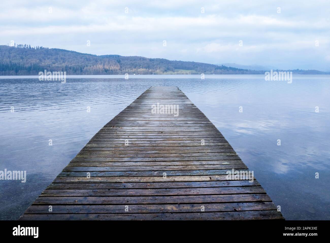Looking down a long wooden jetty with a diminishing persepective, jutting straight out over a clear calm blue lake relecting the white and blue of the Stock Photo