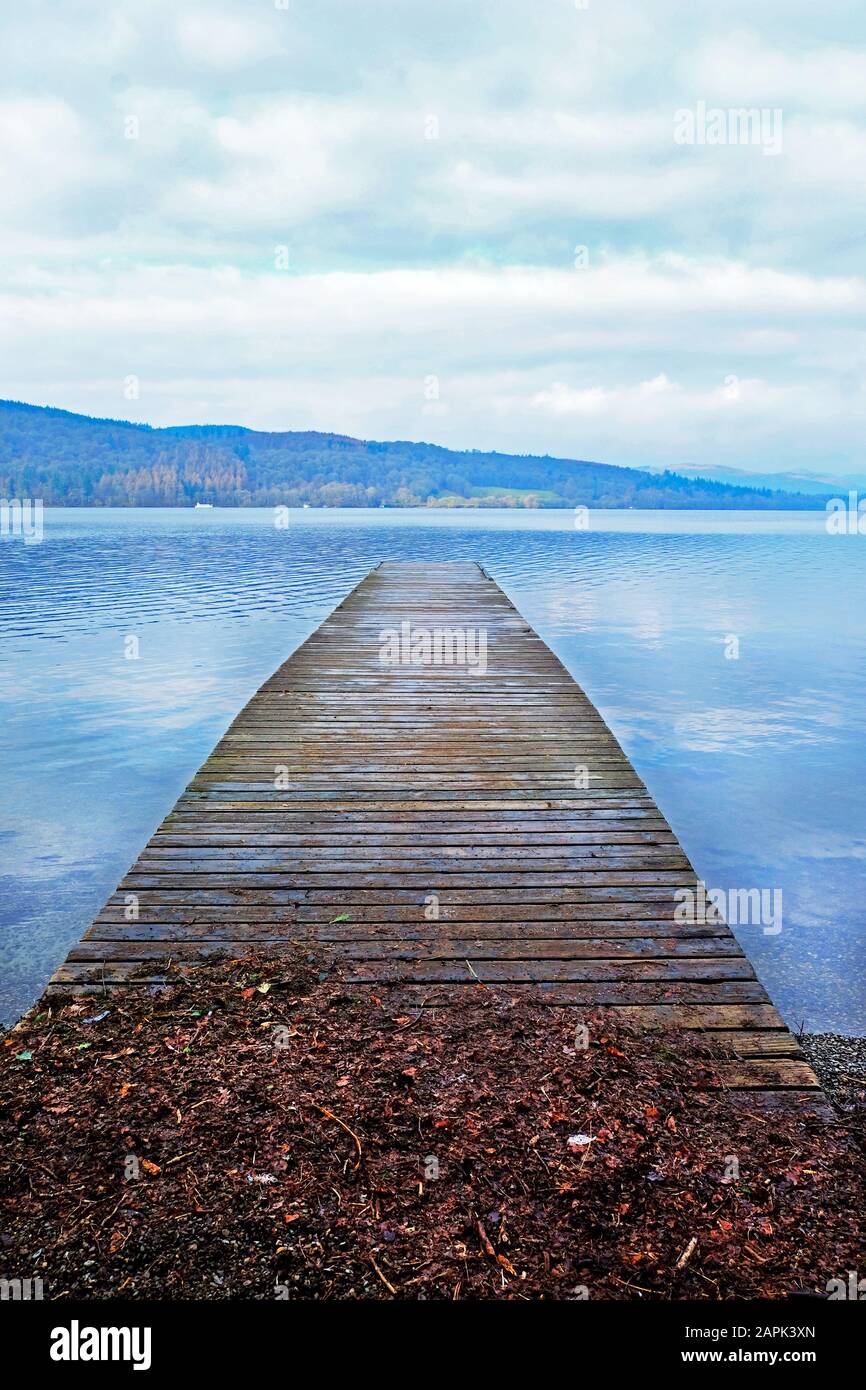 Looking down a long wooden jetty with a diminishing persepective, jutting straight out over a clear calm blue lake relecting the white and blue of the Stock Photo