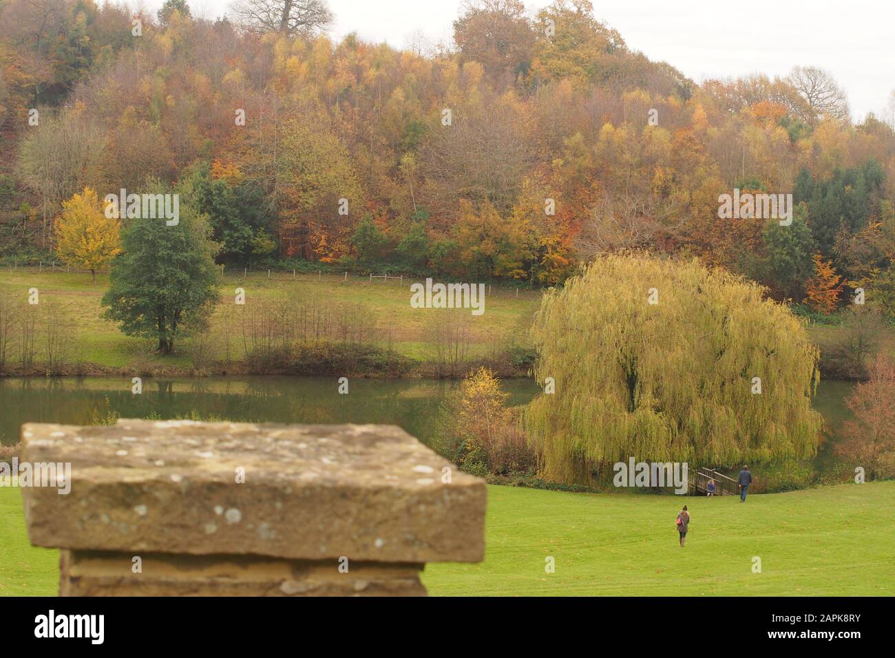A view looking over a stone garden wall to sloping grass with a young family down to a lake with autumn trees and a woodland beyond Stock Photo