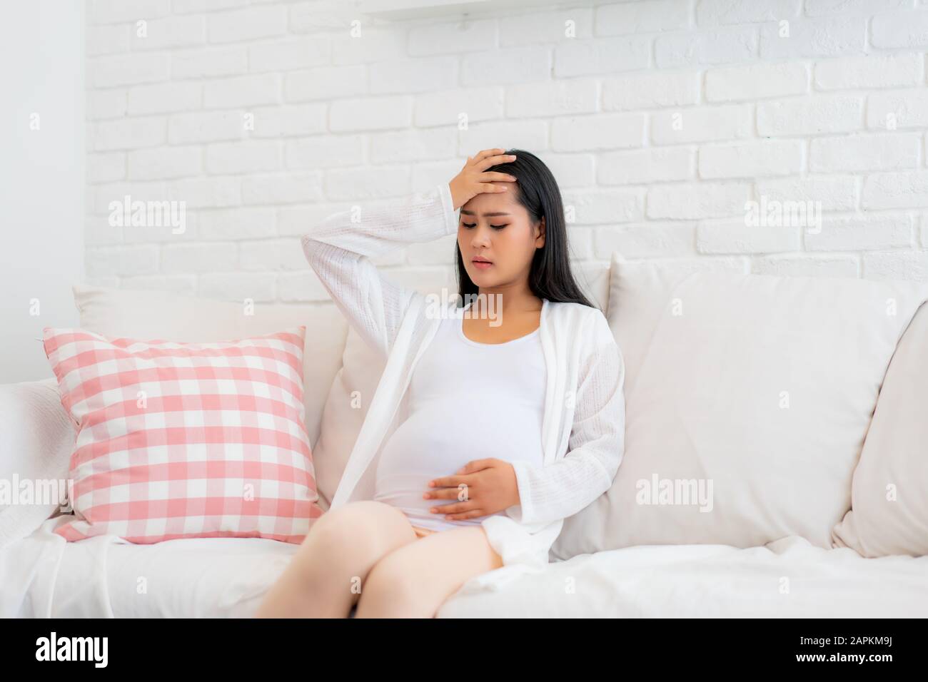 Young pregnant woman with headache sitting on sofa in living room at house. Pregnancy symptoms, expectation, parenthood concept, copy space. Stock Photo