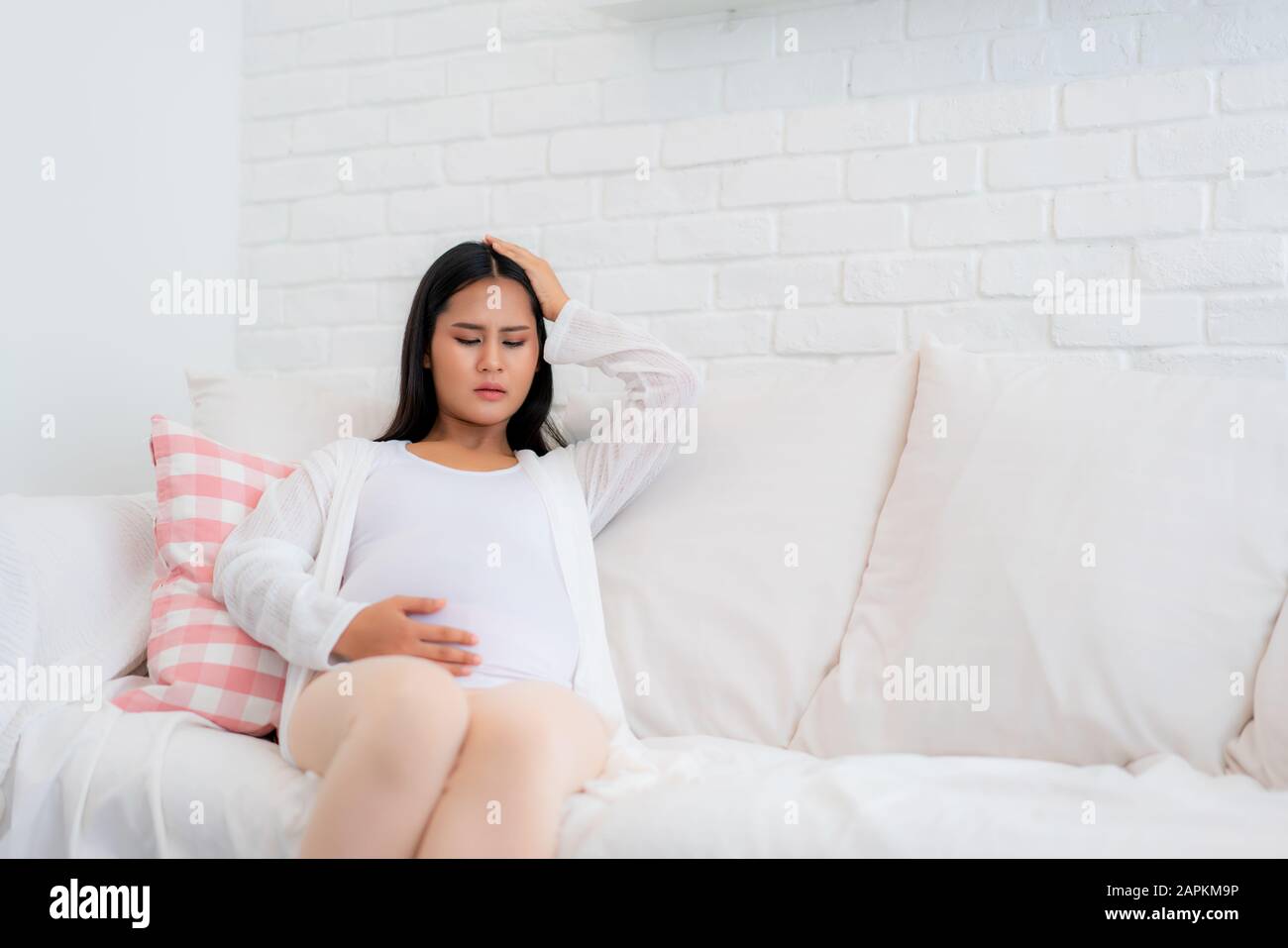 Young pregnant woman with headache sitting on sofa in living room at house. Pregnancy symptoms, expectation, parenthood concept, copy space. Stock Photo