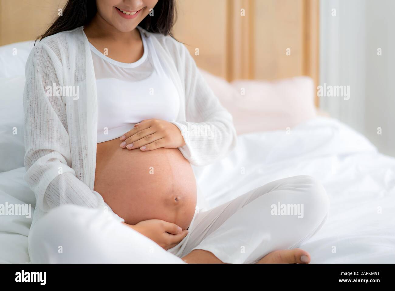 Close up of happy smiling pregnant Asian woman sitting on bed and touching her belly at home. Pregnancy, rest, people and expectation concept Stock Photo