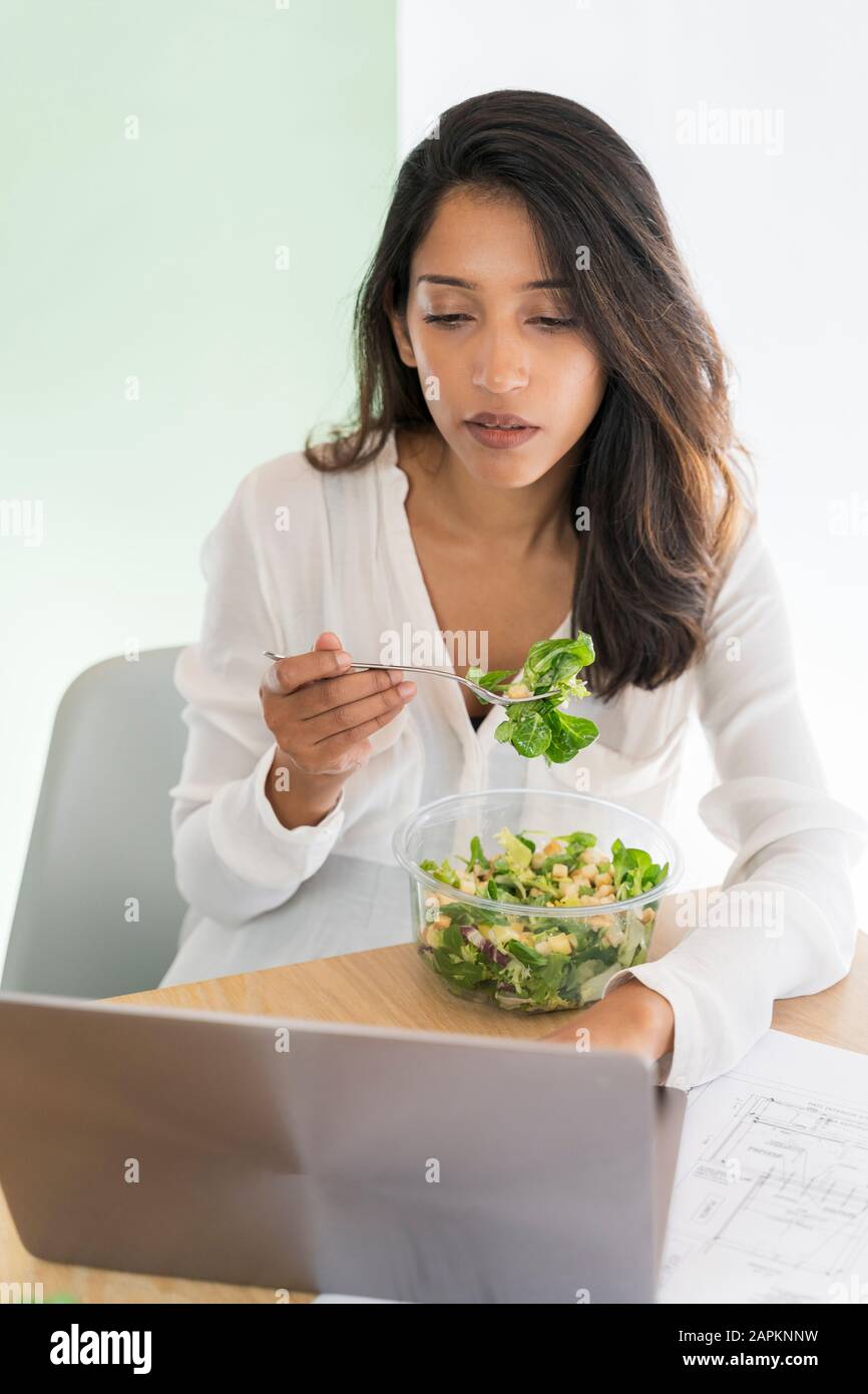 Portrait of young architect eating mixed salad at desk looking at laptop Stock Photo