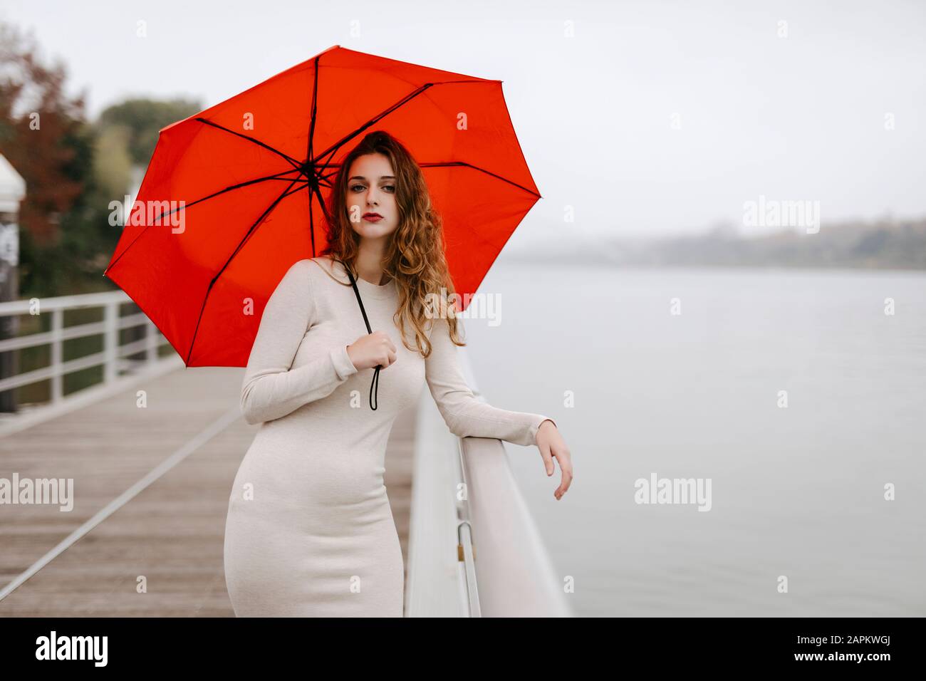 Portrait of young woman with red umbrella, leaning on railing during rainy day Stock Photo