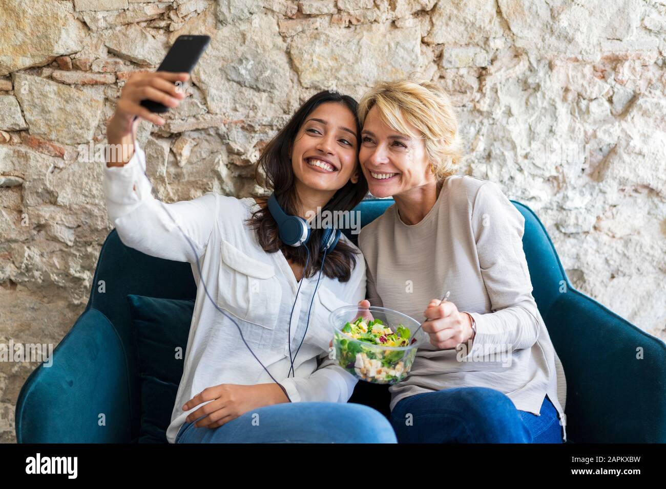 Two happy colleagues sitting on couch at lunchtime taking selfie with smartphone Stock Photo