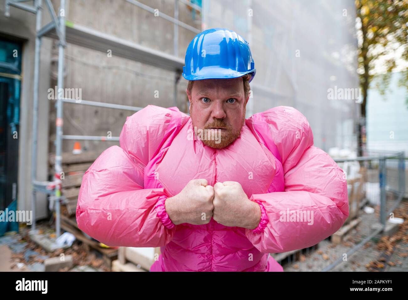 Portrait of man wearing pink bodybuilder costume and hard hat at construction site Stock Photo