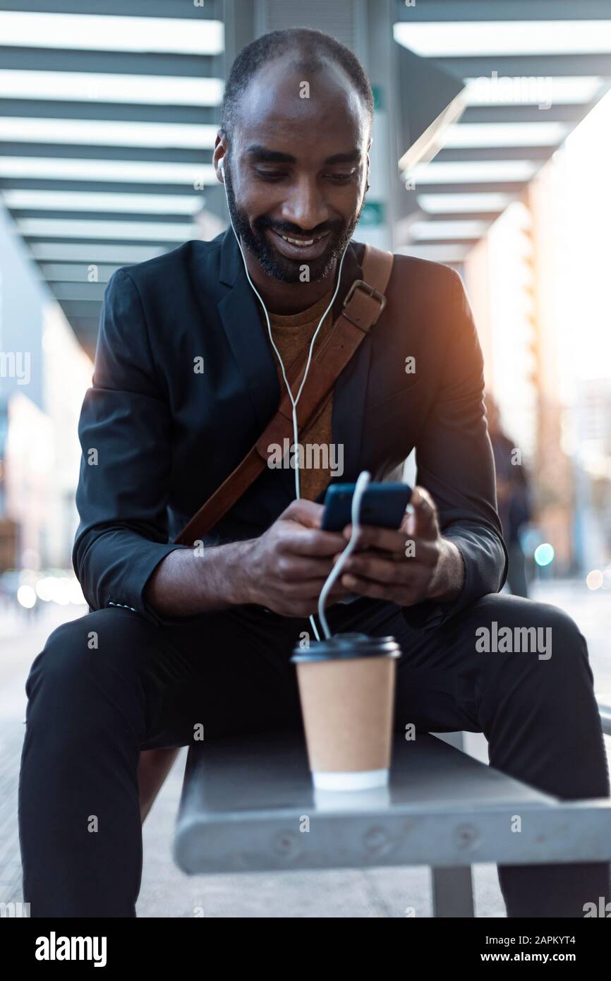 Smiling young businessman sitting at tram stop using earphones and smartphone Stock Photo